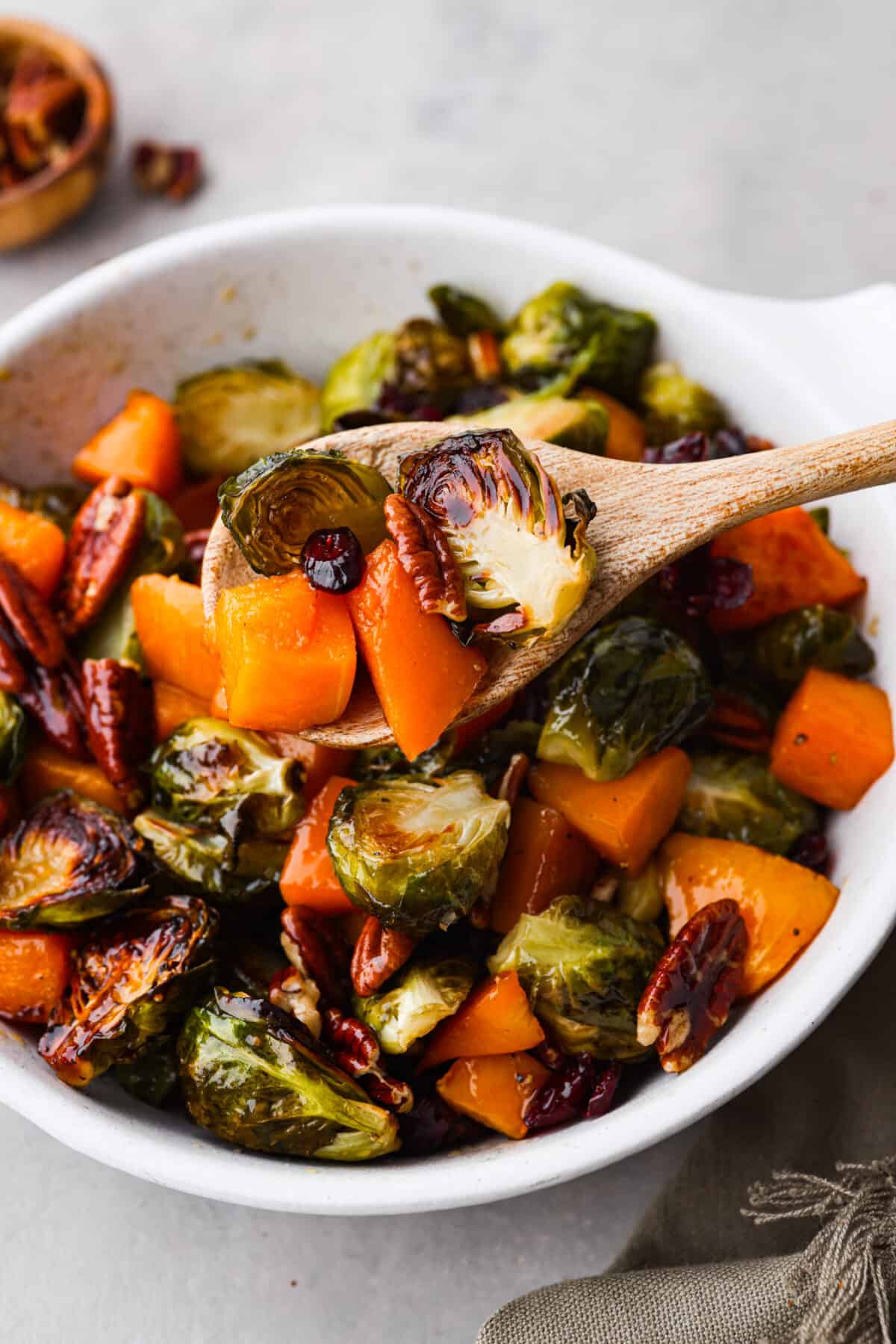 Simple to make and full of incredible flavor, maple roasted brussels sprouts and butternut squash is the perfect side dish. The veggies are crisp on the edges but soft and tender on the inside, tossed in a delicious maple glaze and surrounded by crunchy pecans and cranberries. Maple roasted brussels sprouts and butternut squash in a wooden spoon.