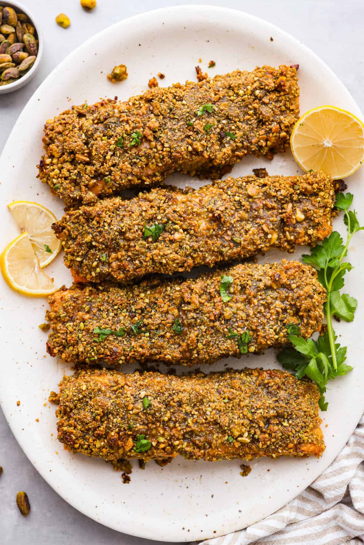 Delicious Pistachio Crusted Salmon is a savory entree that is coated in a mix of garlic, Dijon mustard, honey, and lemon juice with a crunchy pistachio crust all baked to perfection. Ready in just 30 minutes! 4 pistachio crusted salmon filets on a white plate, garnished with lemon.