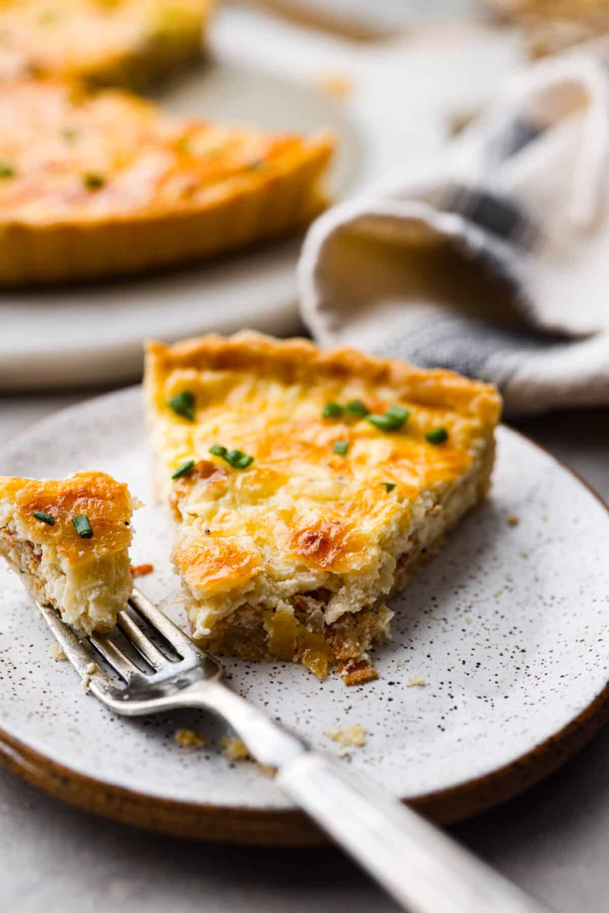 Flavorfulย Quiche Lorraineย is a classic French breakfast recipe. Itโs a simple, yet incredible combination of bacon, shallots, creamy egg custard and cheese, all baked in a flaky pie crust. Perfect for breakfast, brunch, or any meal of the day! A slice of quiche lorraine with a bite taken out of it on a stoneware plate.