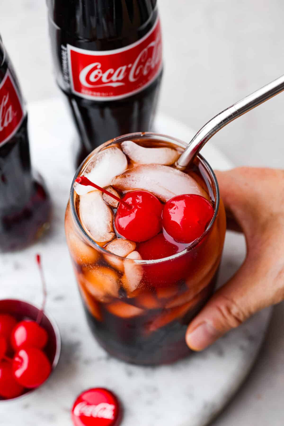 The whole family will love this fun Roy Rogers drink made with Coca-Cola, grenadine syrup, and Maraschino cherries on top! Only 3 simple ingredients to enjoy this classic non-alcoholic drink. Top view of a hand holding a drink with a straw and cherries on top.