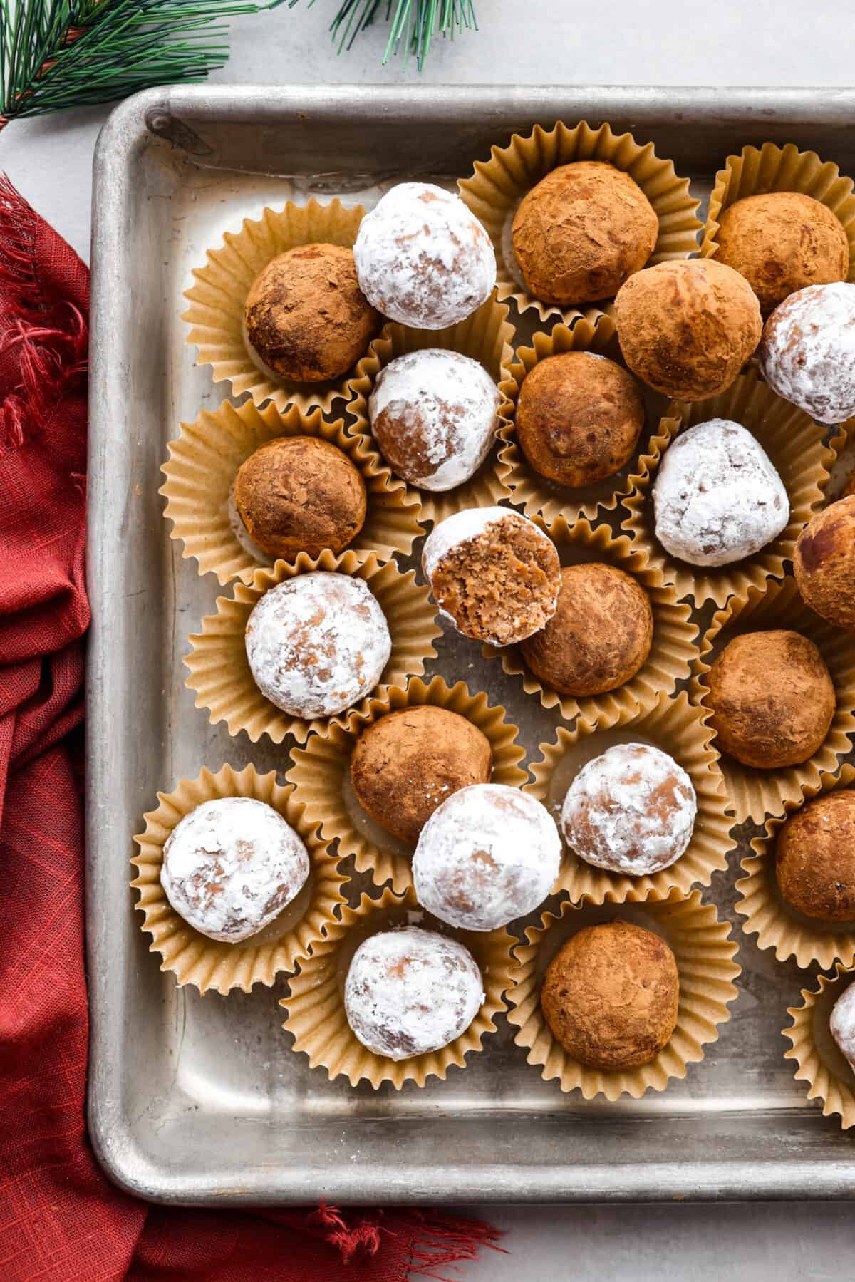 Easy and impressive rum balls are perfect for the holidays! Vanilla wafer crumbs, powdered sugar, cocoa powder, and rum create a truffle-like consistency bursting with rich flavors. A simple bite-sized dessert that every party needs! Top close view of rum balls in mini muffin cups and stacked on a sheet pan.