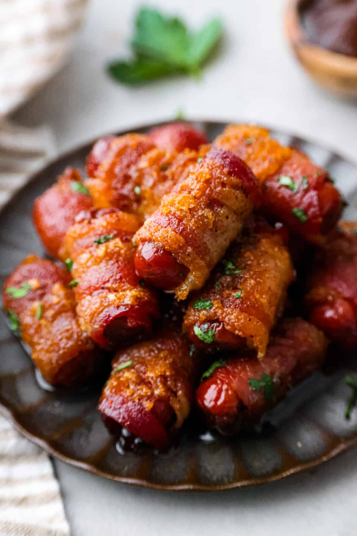 These bacon wrapped smokies are the perfect sweet and salty appetizer that everyone will devour! Little smokie sausagesย are wrapped in crispy bacon and caramelized in a brown sugar spice blend. They are sure to be a favorite at your next party! Close view of bacon wrapped smokies stacked on a brown plate.