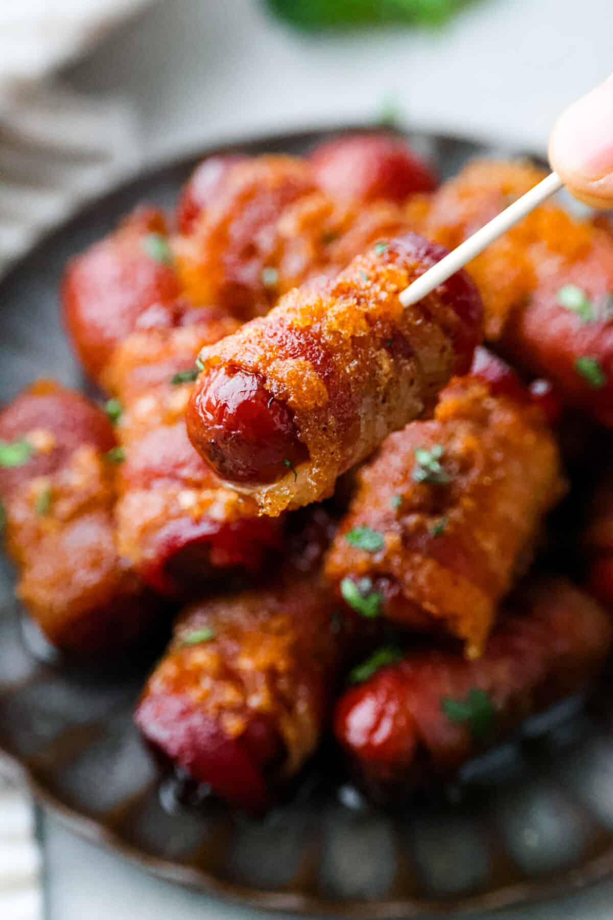 These bacon wrapped smokies are the perfect sweet and salty appetizer that everyone will devour! Little smokie sausagesย are wrapped in crispy bacon and caramelized in a brown sugar spice blend. They are sure to be a favorite at your next party! Close view of a toothpick holding a bacon wrapped smokie.