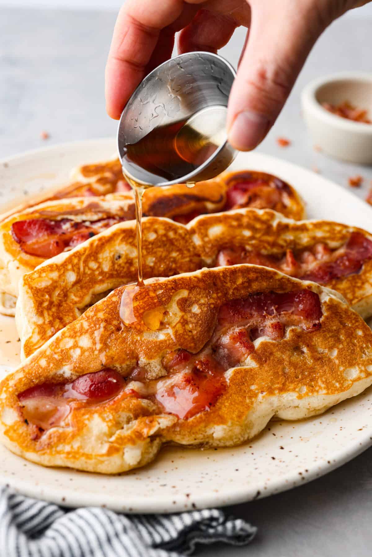 An all-in-one breakfast, and so good, you won't be able to get enough. Crispy bacon nestled in pillowy pancakes, topped with butter and syrup - what could be better? Pouring maple syrup over bacon pancakes.