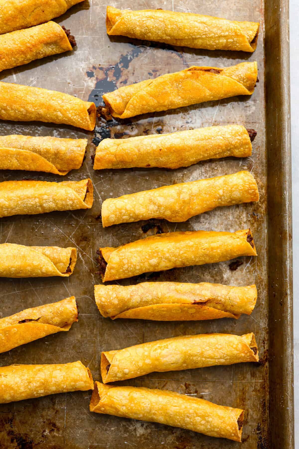 Cook up a batch of these homemade beef taquitos, you won't be disappointed! The meaty, cheesy filling and crispy tortilla shell are to die for. A million times better than the frozen kind! Baked beef taquitos on a baking sheet.