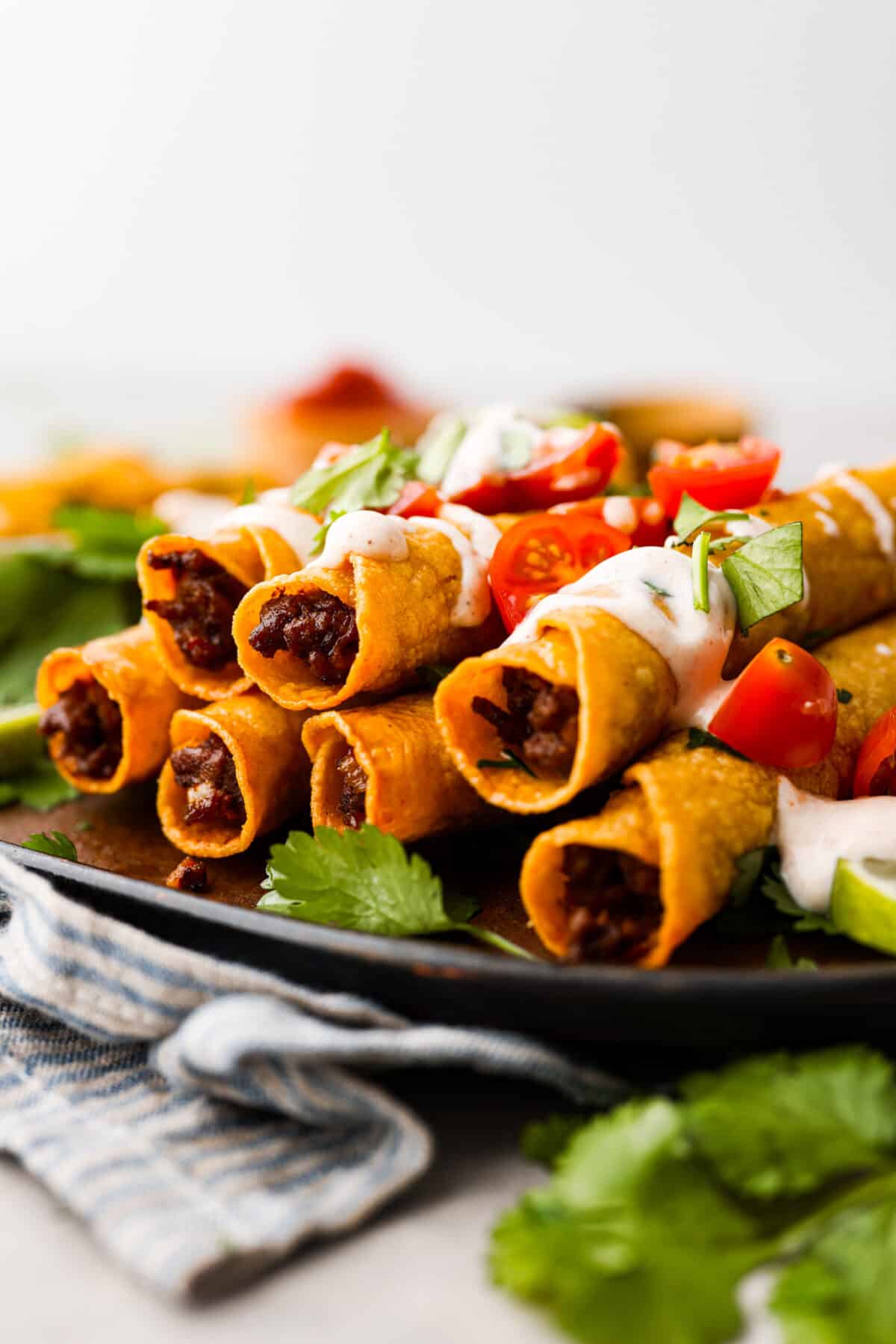 Cook up a batch of these homemade beef taquitos, you won't be disappointed! The meaty, cheesy filling and crispy tortilla shell are to die for. A million times better than the frozen kind! Beef taquitos on a black plate, topped with chopped tomatoes and sour cream.