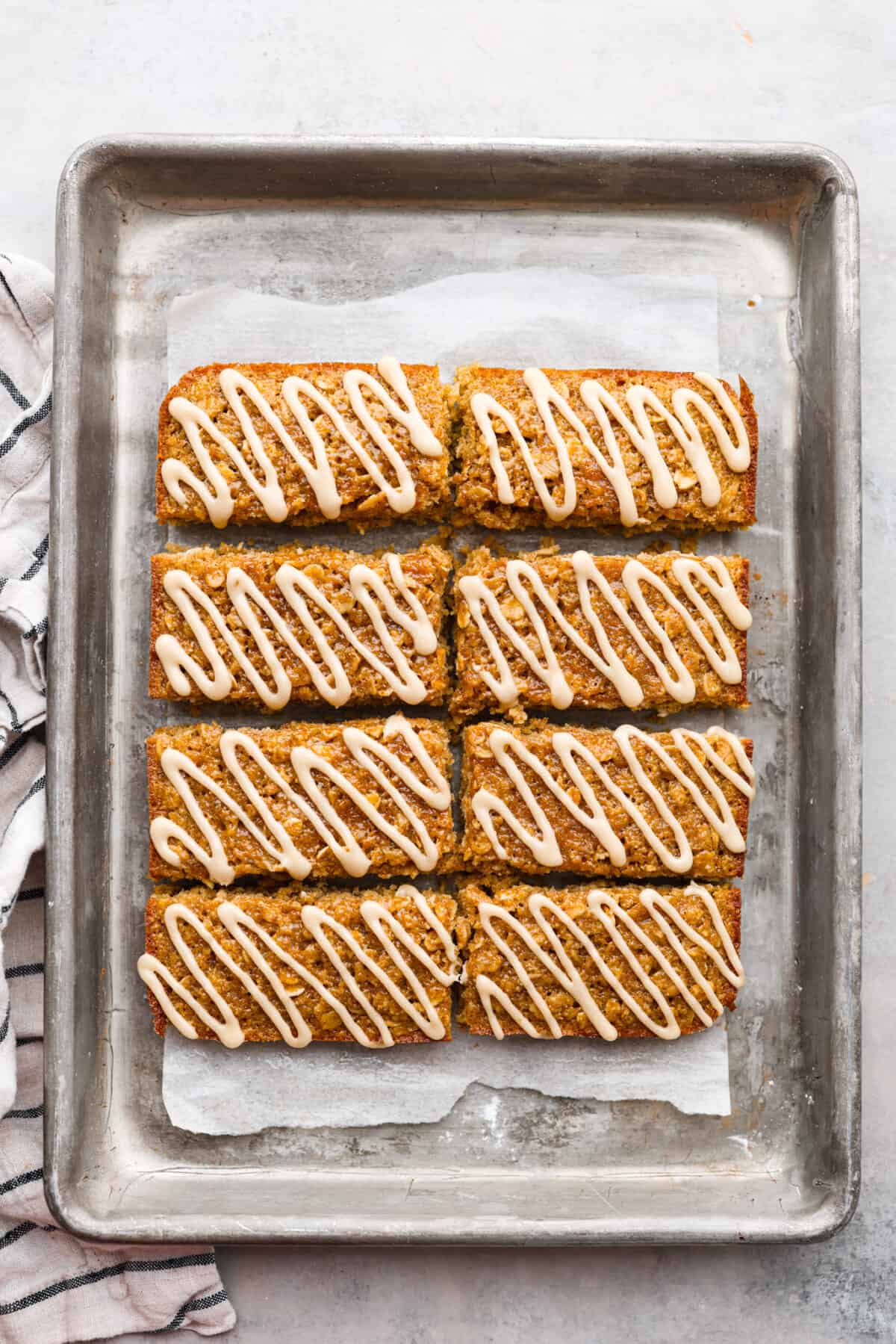 Start your day right with these simple and delicious Oatmeal Breakfast Bars. Topped with a sweet brown sugar maple glaze, these bars are easy to whip up and guaranteed to be a hit for a wholesome morning treat. Cooked breakfast bars with a glaze that are on parchment paper on a baking sheet.