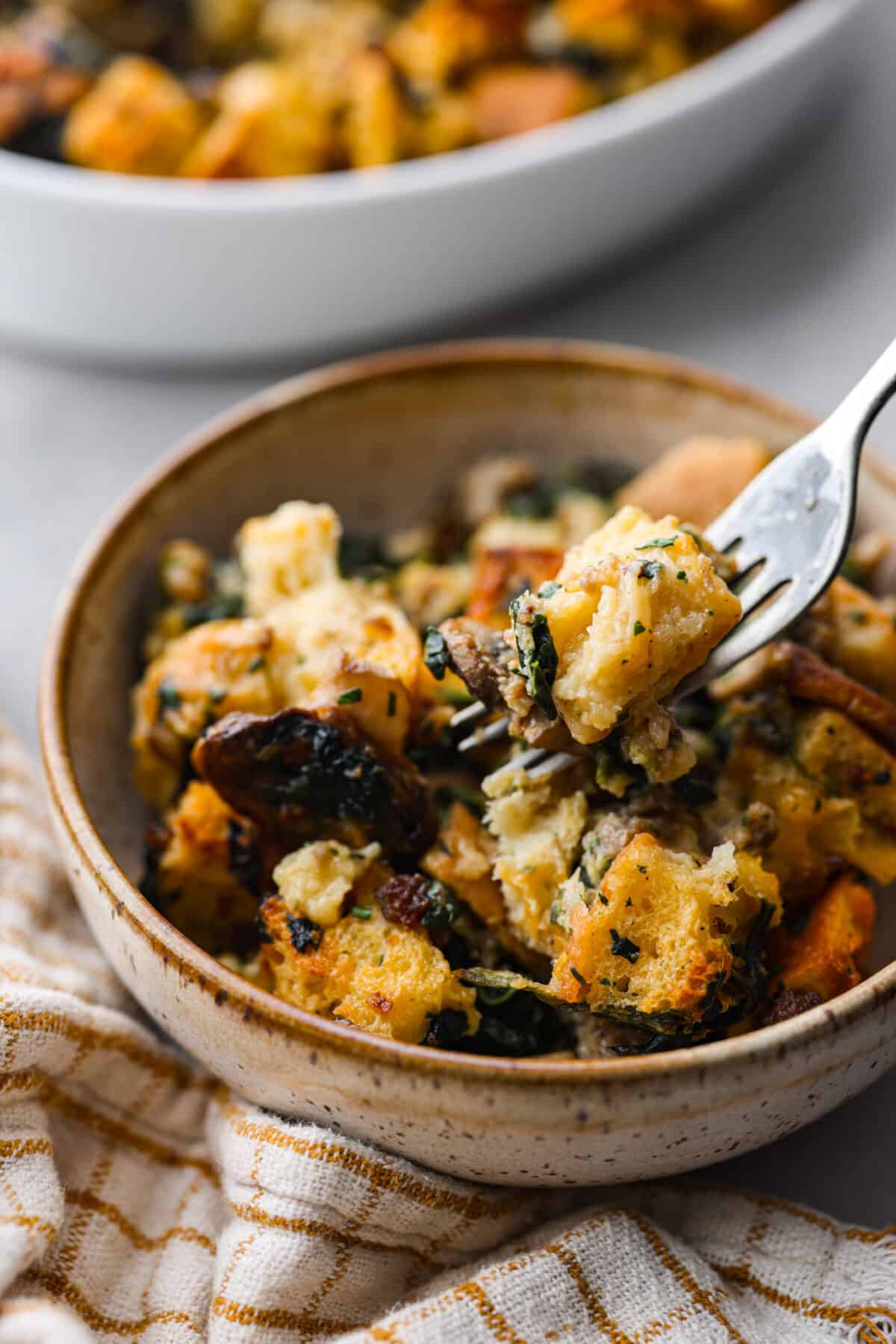 This breakfast strata is bursting with savory sausage, tender mushrooms, soft bread pieces, and melty cheddar. Perfect for your next holiday or family breakfast. It is packed with so much delicious flavor and texture that it will leave you wanting seconds! Angle shot of plated breakfast strata in a bowl.