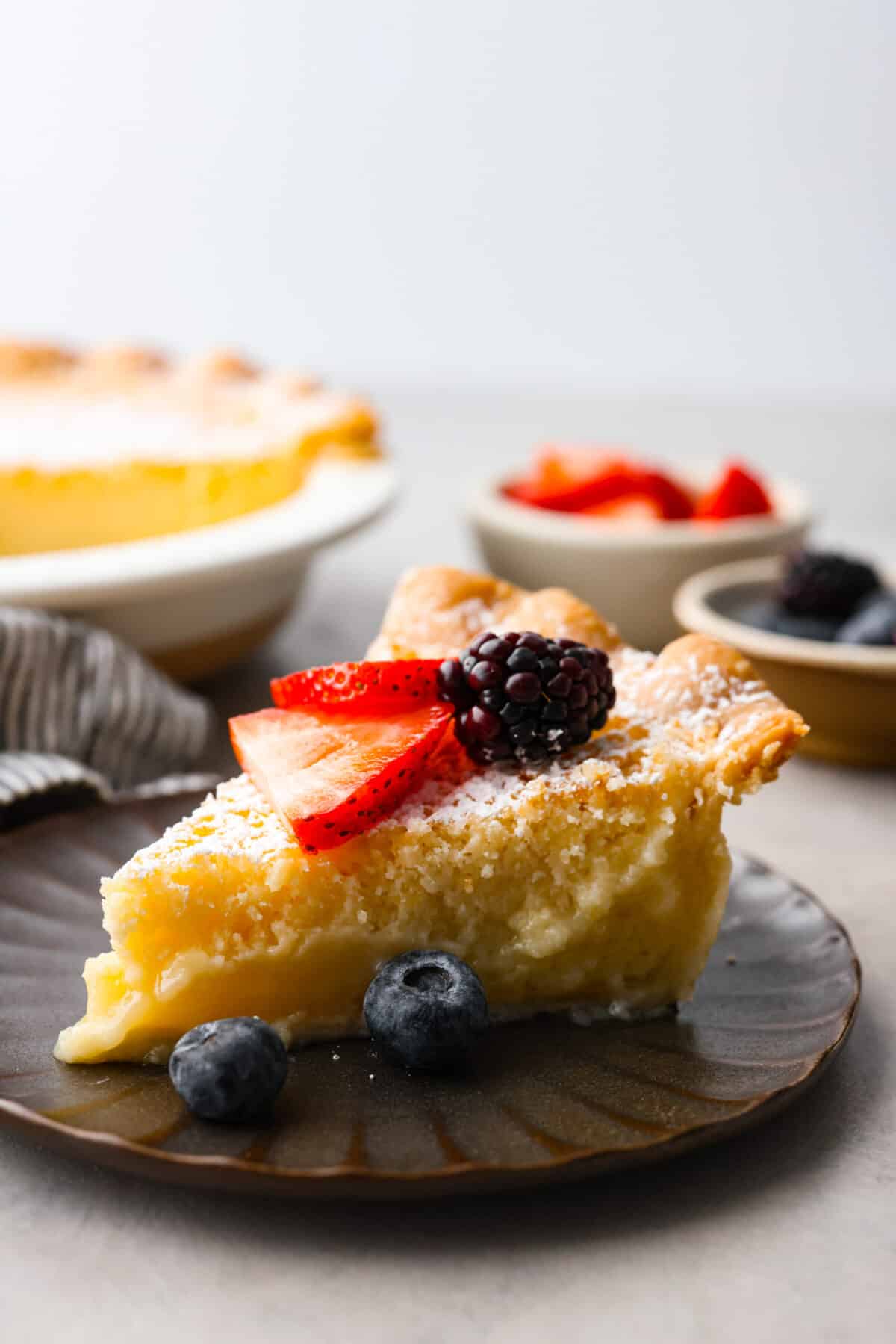 This amazing buttermilk pie is made with a creamy, lemon-kissed custard tucked inside a buttery, golden crust. So delicious and perfect for any time of year. It's like sunshine in pie form! A slice of pie topped with strawberries, blackberries, and blueberries