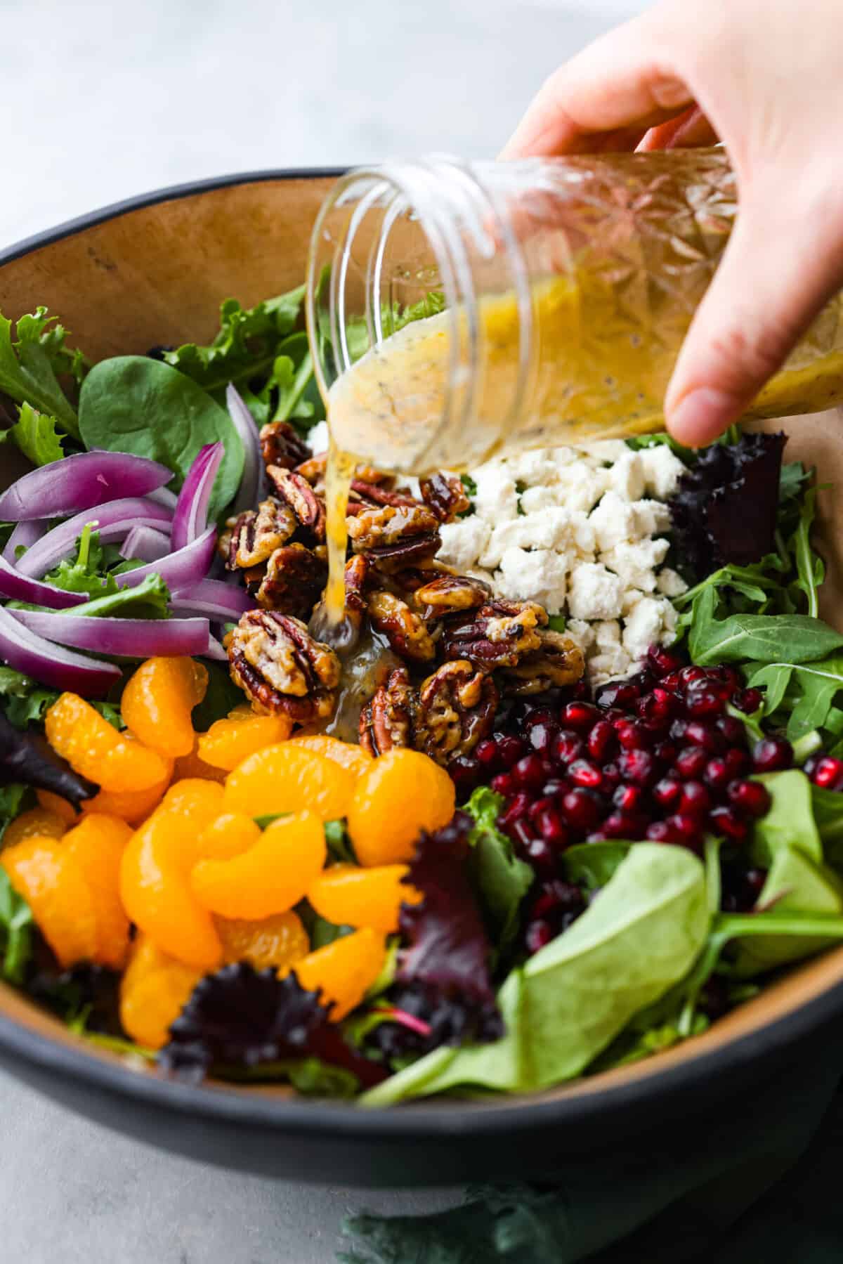 Serve this refreshing festive Christmas salad with your holiday menu! It's a beautiful wintery salad full of mixed greens, orange slices, juicy pomegranates, red onion, candied pecans, and crumbled feta cheese. All tossed together in an orange vinaigrette. Close side view of the vinaigrette pouring over the salad ingredients.