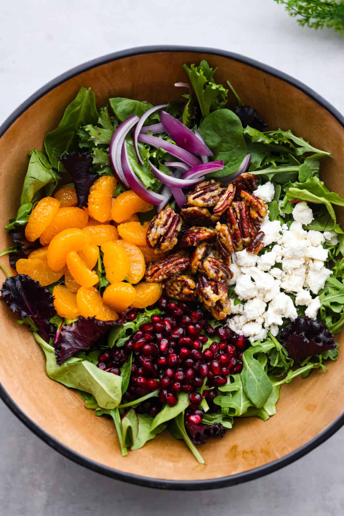 Serve this refreshing festive Christmas salad with your holiday menu! It's a beautiful wintery salad full of mixed greens, orange slices, juicy pomegranates, red onion, candied pecans, and crumbled feta cheese. All tossed together in an orange vinaigrette. Top view of salad ingredients added to a bowl.