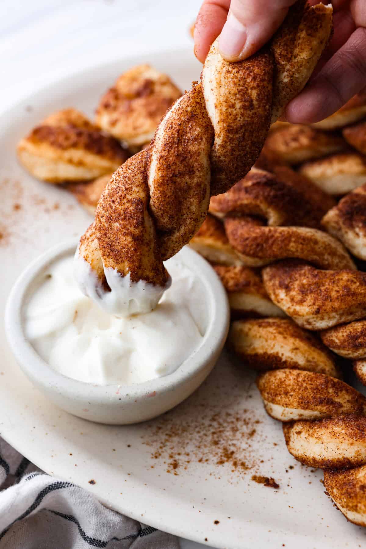 Everyone LOVES these sweet and fluffy cinnamon roll breadtwists. Homemade dough twisted and baked in brown sugar cinnamon goodness. It's like eating the middle of a cinnamon roll! Perfect for breakfast, enjoy as a snack, or delicious as a dessert. Close view of a cinnamon bread twist dipping in frosting.