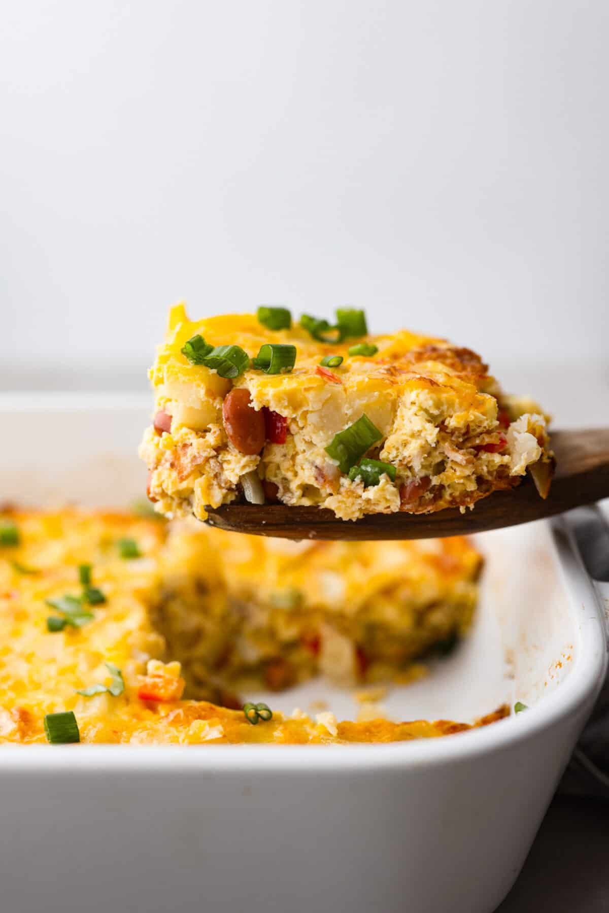 Cowboy breakfast casserole is a hearty dish that's chock full of breakfast favorites (eggs, bacon, sausage, beans, cheese, potatoes, and more!) and super easy to make! A slice of breakfast casserole being lifted out of the pan with a wooden spoon.
