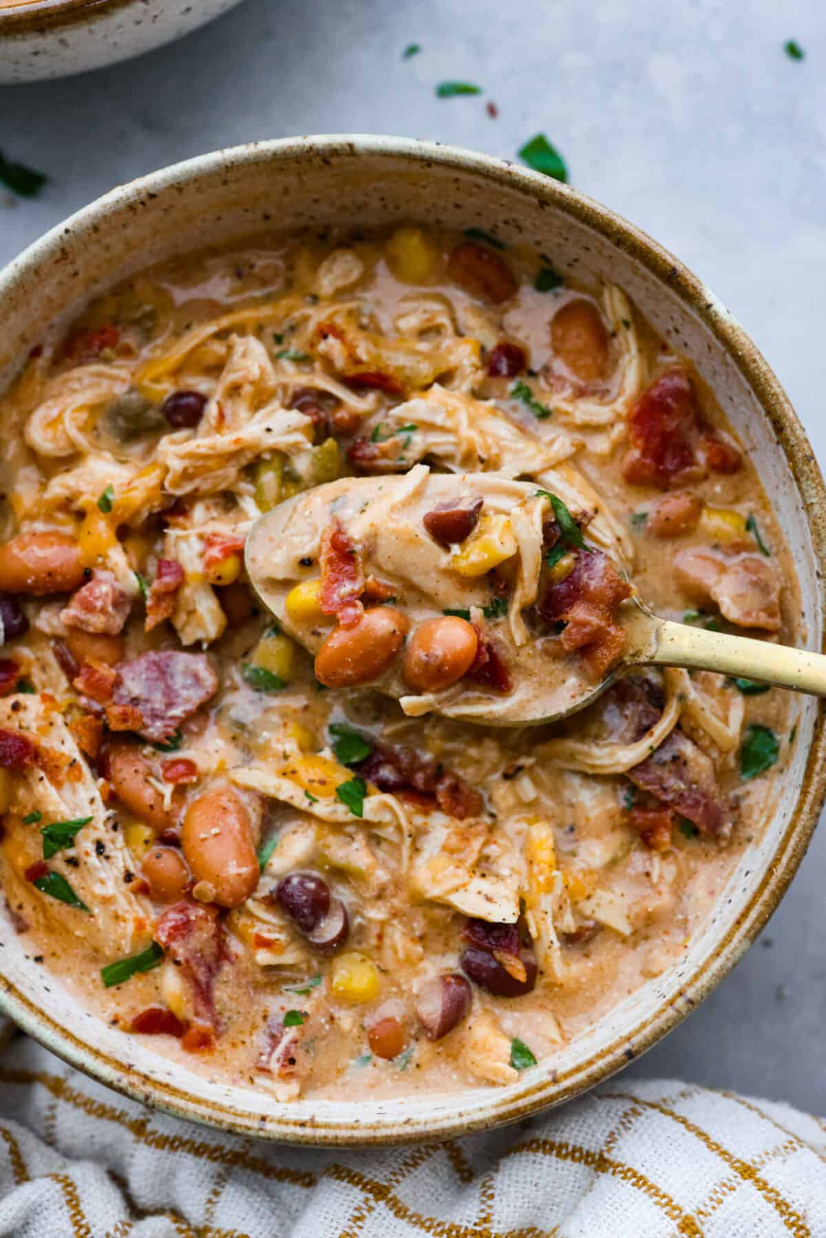 Creamy crack chicken chili is an explosion of irresistible flavors! It's loaded with shredded chicken, tender beans, corn, AND all your favorite crack-inspired ingredients. This is our new favorite chili recipe and we can't get enough of it! Close view of a spoon lifting chili from a bowl.