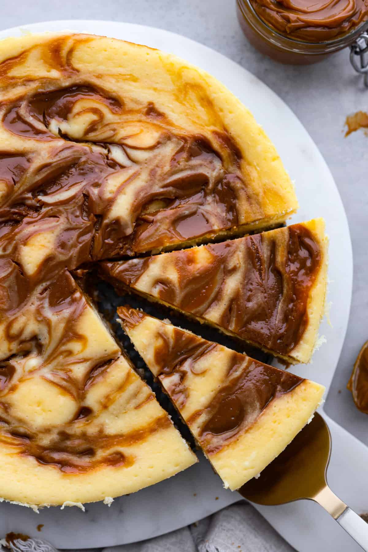 Dulce de leche cheesecake is the most creamy and decadent dessert, with swirls of caramel in every bite. It's guaranteed to be the star of your dinner table! Top-down view of dulce de leche cheesecake, cut into slices and being served with a metal cake spatula.