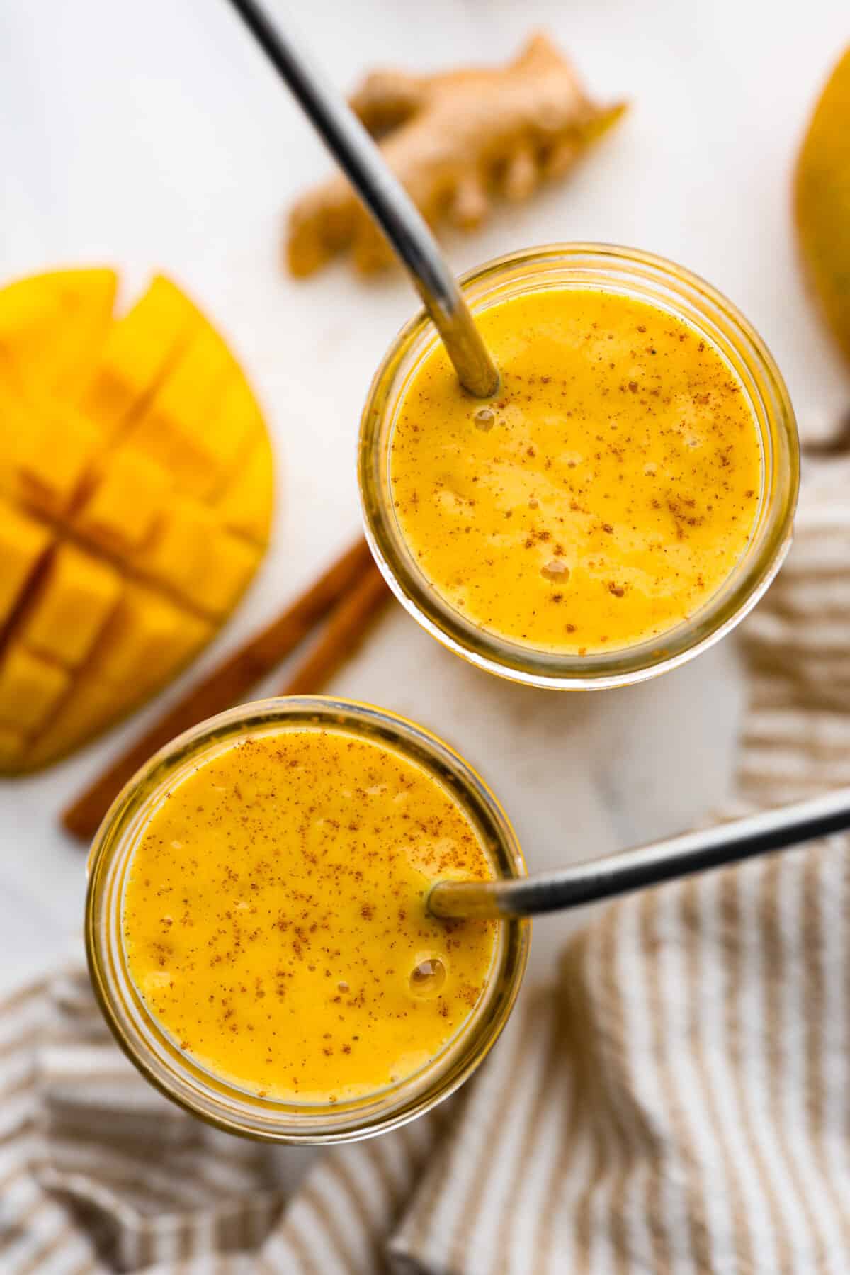 This creamy and sweet golden milk smoothie will brighten your morning! The smooth texture comes from the mangos, Greek yogurt, and banana. It's spiced to perfection and bursting with healthy benefits. Top view of the smoothie in two glasses. Each glass has a metal straw and cinnamon garnished on top.