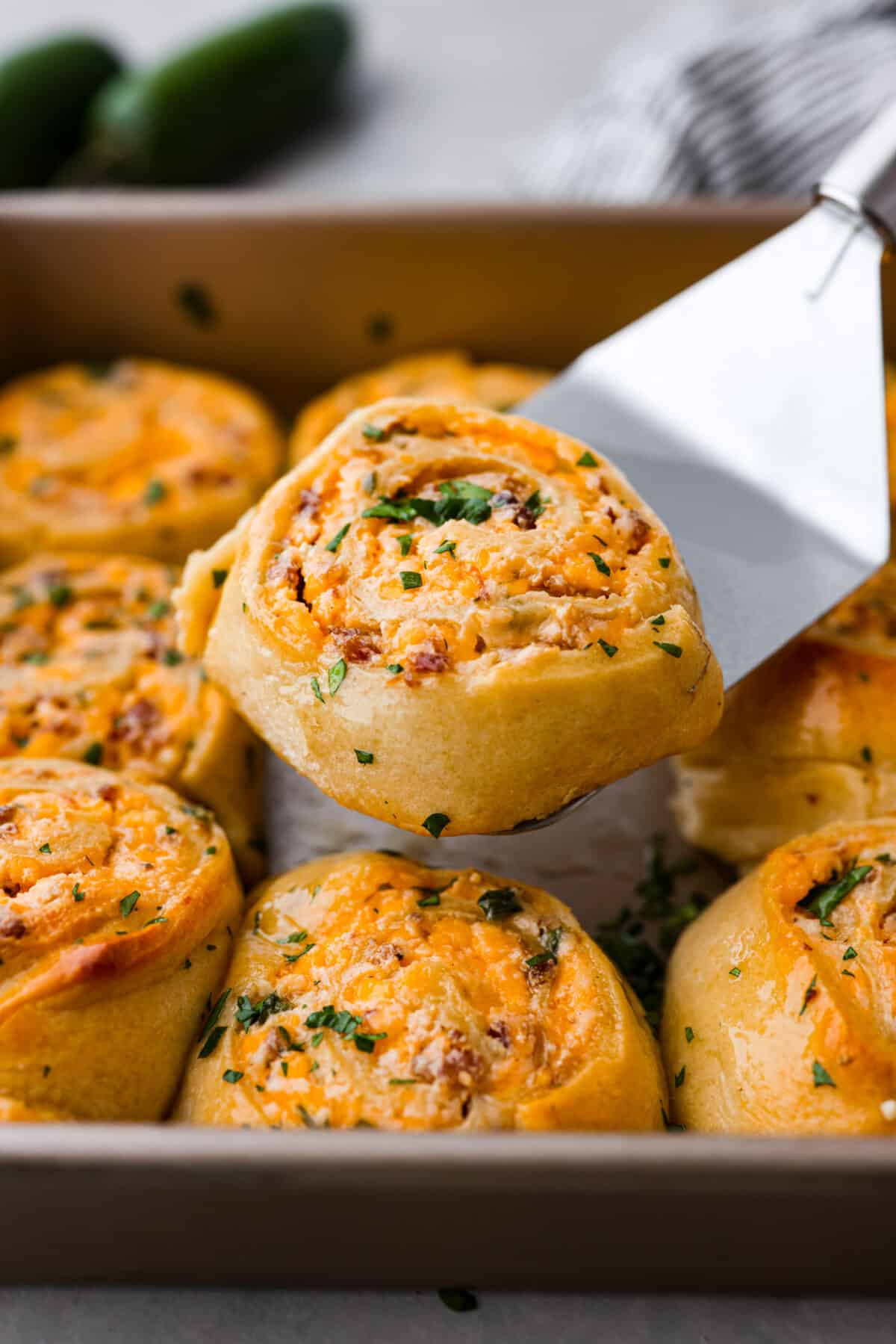 These jalapeño popper rolls are filled with a cream cheese mixture including bacon, cheese, and diced jalapeño and rolled up in crescent dough. Baked then brushed with garlic butter...everyone will devour them! Close view of a spatula lifting up a jalapeno popper roll from a pan.