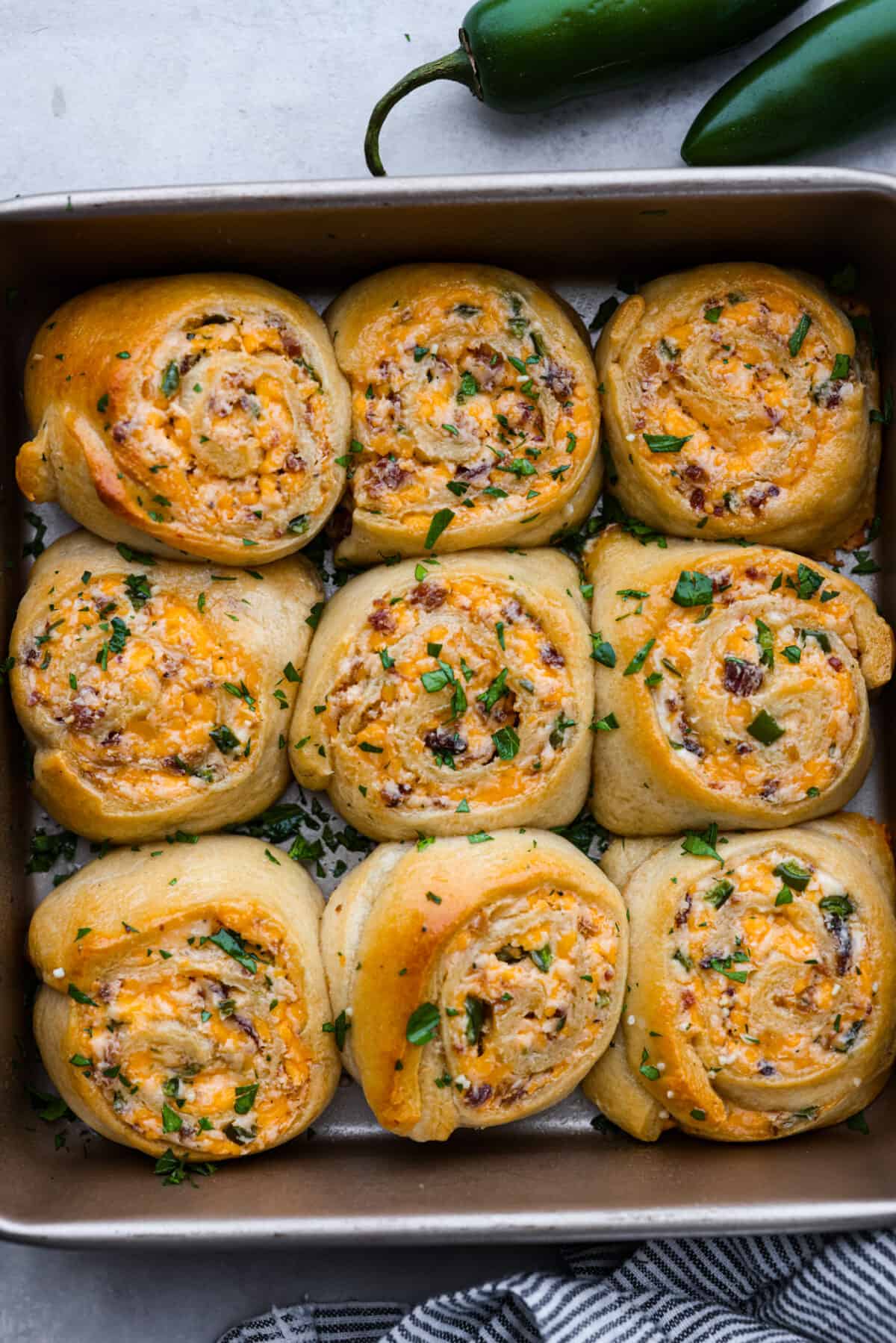 These jalapeño popper rolls are filled with a cream cheese mixture including bacon, cheese, and diced jalapeño and rolled up in crescent dough. Baked then brushed with garlic butter...everyone will devour them! Top view of jalapeno popper rolls baked in a pan.