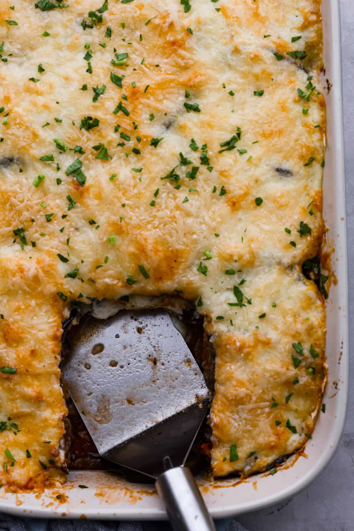 Moussaka is a hearty, delicious dish perfect for a special occasion or a weeknight meal. If you've never tried it before, you're in for a treat! It's made with layers of eggplant, potatoes, meat sauce, and bechamel sauce, and baked until the crust is golden brown. Everyone at the dinner table will be blown away! Top-down view of moussaka in a baking dish with a slice taken out of it.