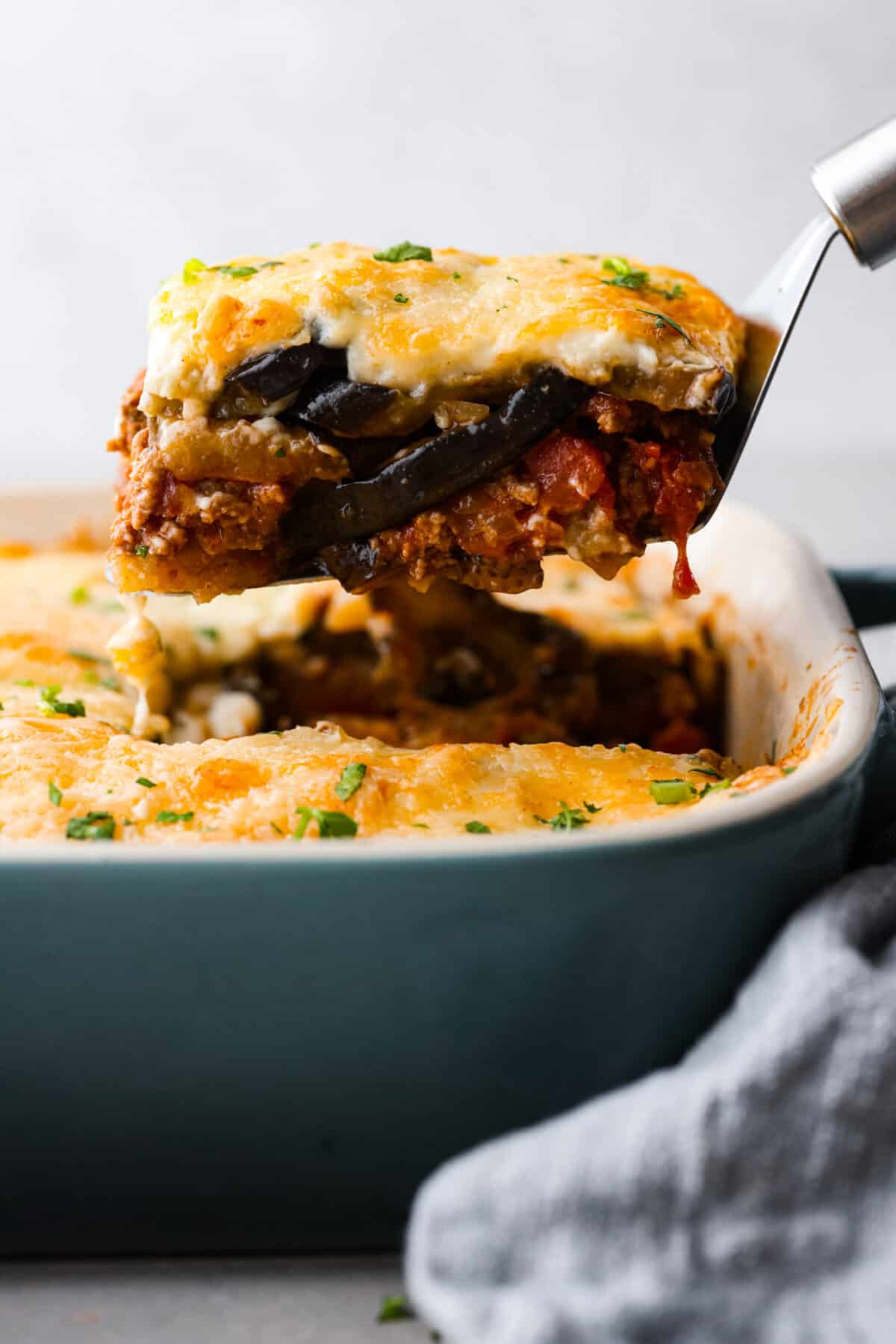Moussaka is a hearty, delicious dish perfect for a special occasion or a weeknight meal. If you've never tried it before, you're in for a treat! It's made with layers of eggplant, potatoes, meat sauce, and bechamel sauce, and baked until the crust is golden brown. Everyone at the dinner table will be blown away! A slice of moussaka being served.