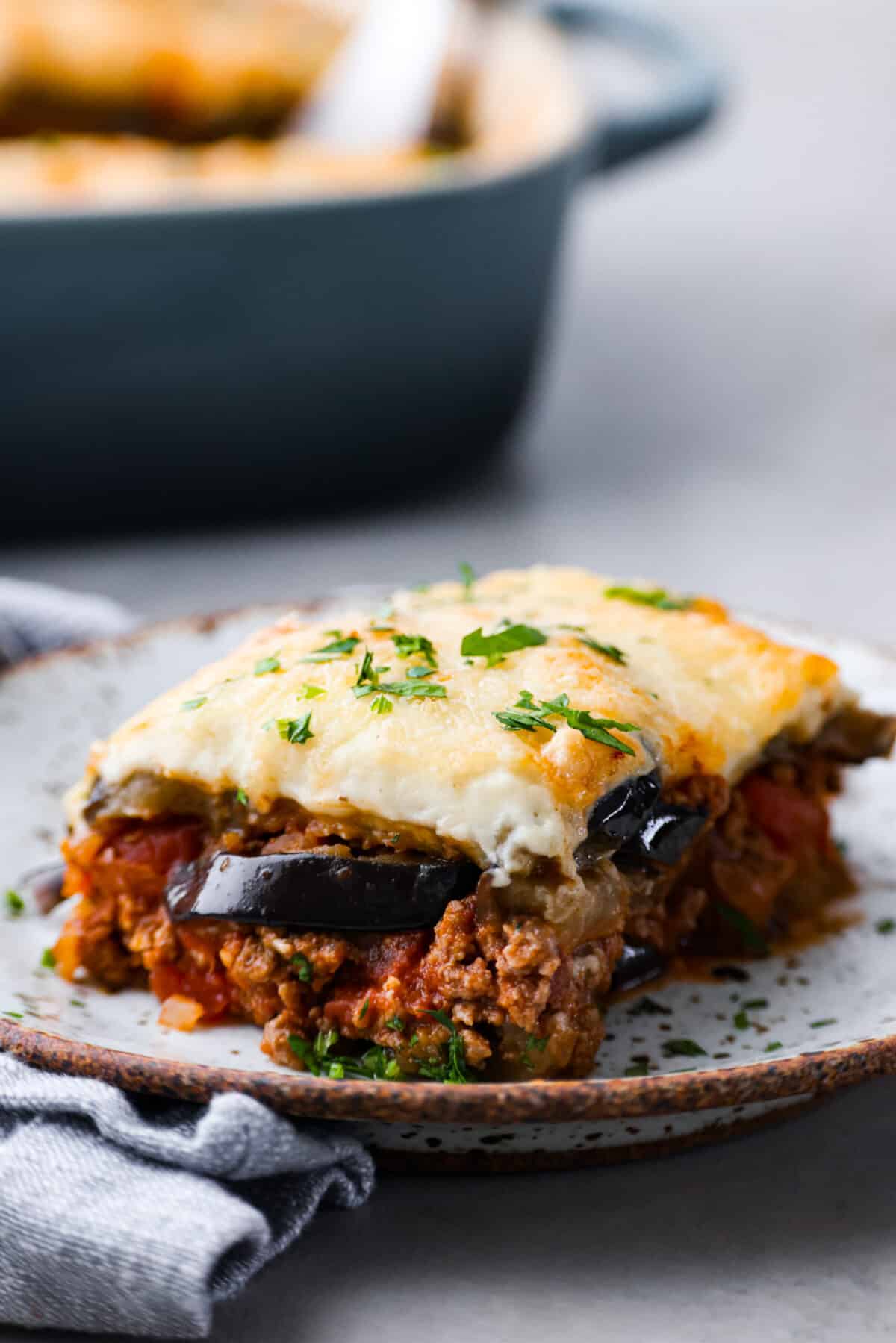 Moussaka is a hearty, delicious dish perfect for a special occasion or a weeknight meal. If you've never tried it before, you're in for a treat! It's made with layers of eggplant, potatoes, meat sauce, and bechamel sauce, and baked until the crust is golden brown. Everyone at the dinner table will be blown away! A slice of moussaka on a stoneware plate.