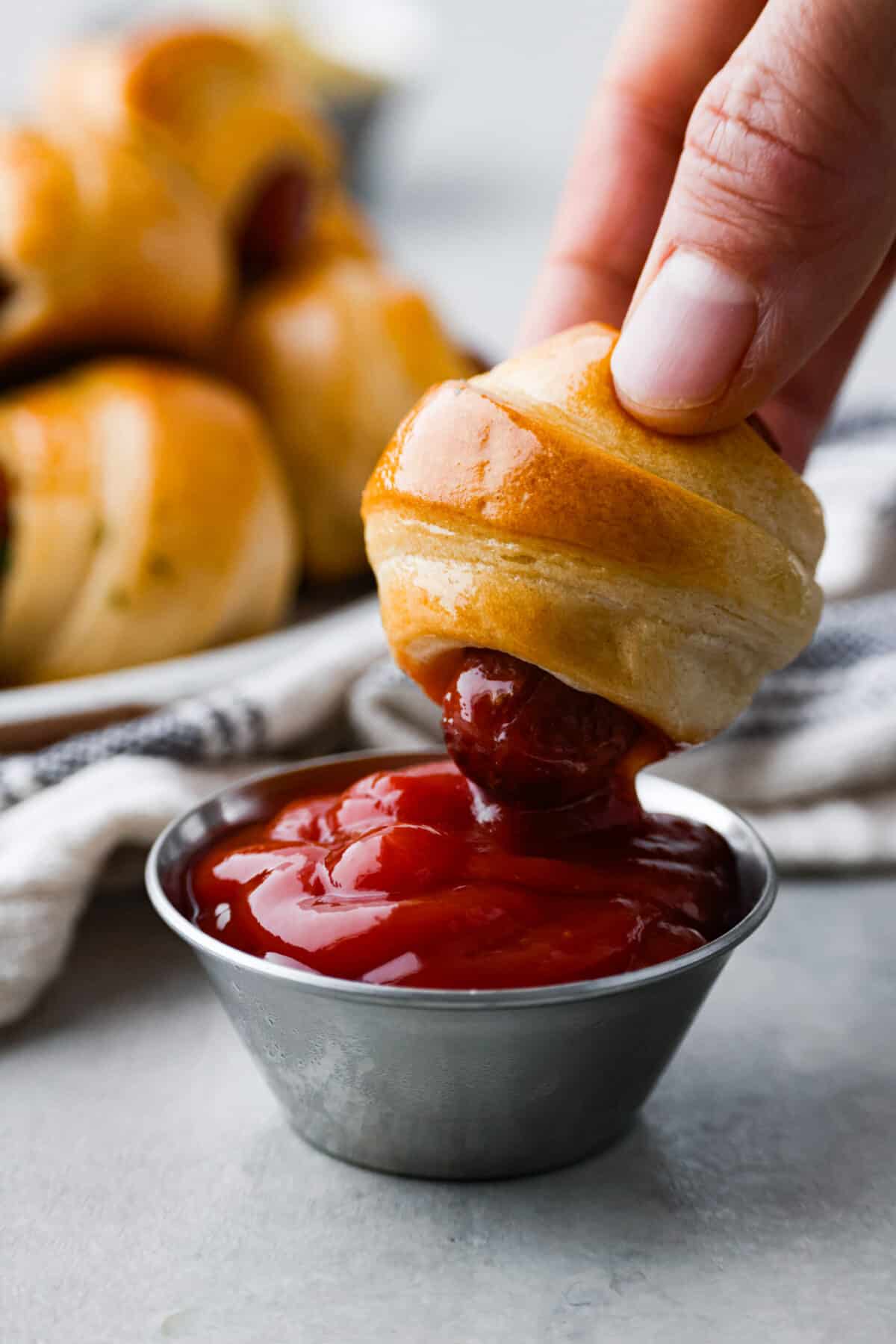 Gotta love a classic Pigs in a Blanket appetizer! These are always the first to go at a party, and for good reason. Whole all-beef hot dogs are wrapped in crescent roll dough, stuffed with cheeses and baked with a garlicky butter. They're cheesy, easy to make, and so addictive! Closeup of one of them being dipped into ketchup.