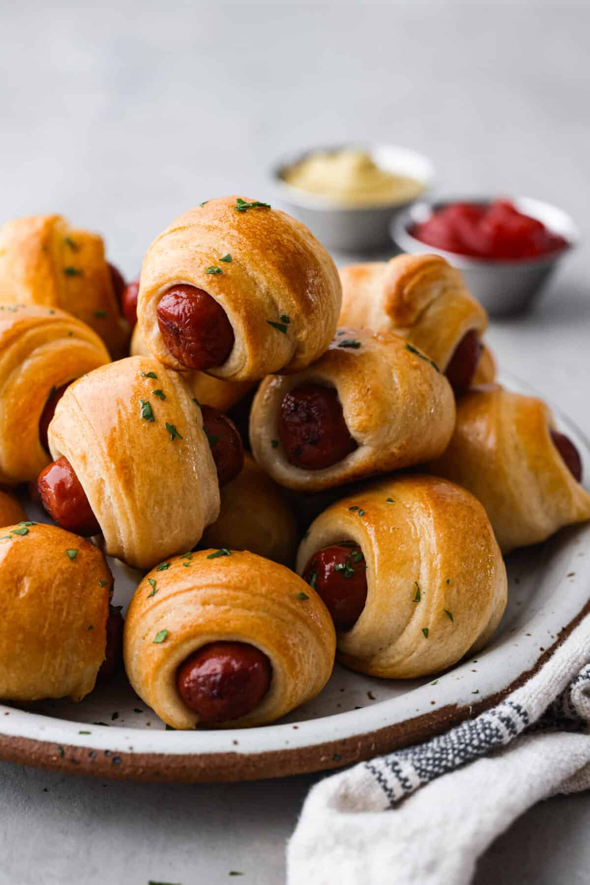 Gotta love a classic Pigs in a Blanket appetizer! These are always the first to go at a party, and for good reason. Whole all-beef hot dogs are wrapped in crescent roll dough, stuffed with cheeses and baked with a garlicky butter. They're cheesy, easy to make, and so addictive! A stack of pigs in a blanket on a stoneware plate.