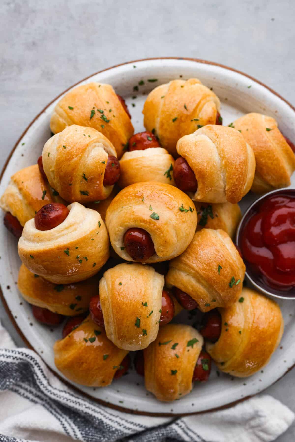 Gotta love a classic Pigs in a Blanket appetizer! These are always the first to go at a party, and for good reason. Whole all-beef hot dogs are wrapped in crescent roll dough, stuffed with cheeses and baked with a garlicky butter. They're cheesy, easy to make, and so addictive! Top-down view of pigs in a blanket served with ketchup.