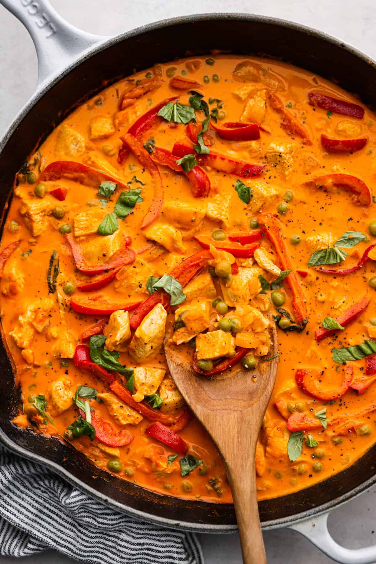 Mouthwatering Red Curry is an explosion of flavors that you're going to love! A combination of tender chicken, colorful red bell peppers, and aromatic Thai basil come together in a creamy coconut milk and curry paste sauce, for an incredibly delicious meal. Top-down view of red curry in a skillet, being stirred with a wooden spoon.