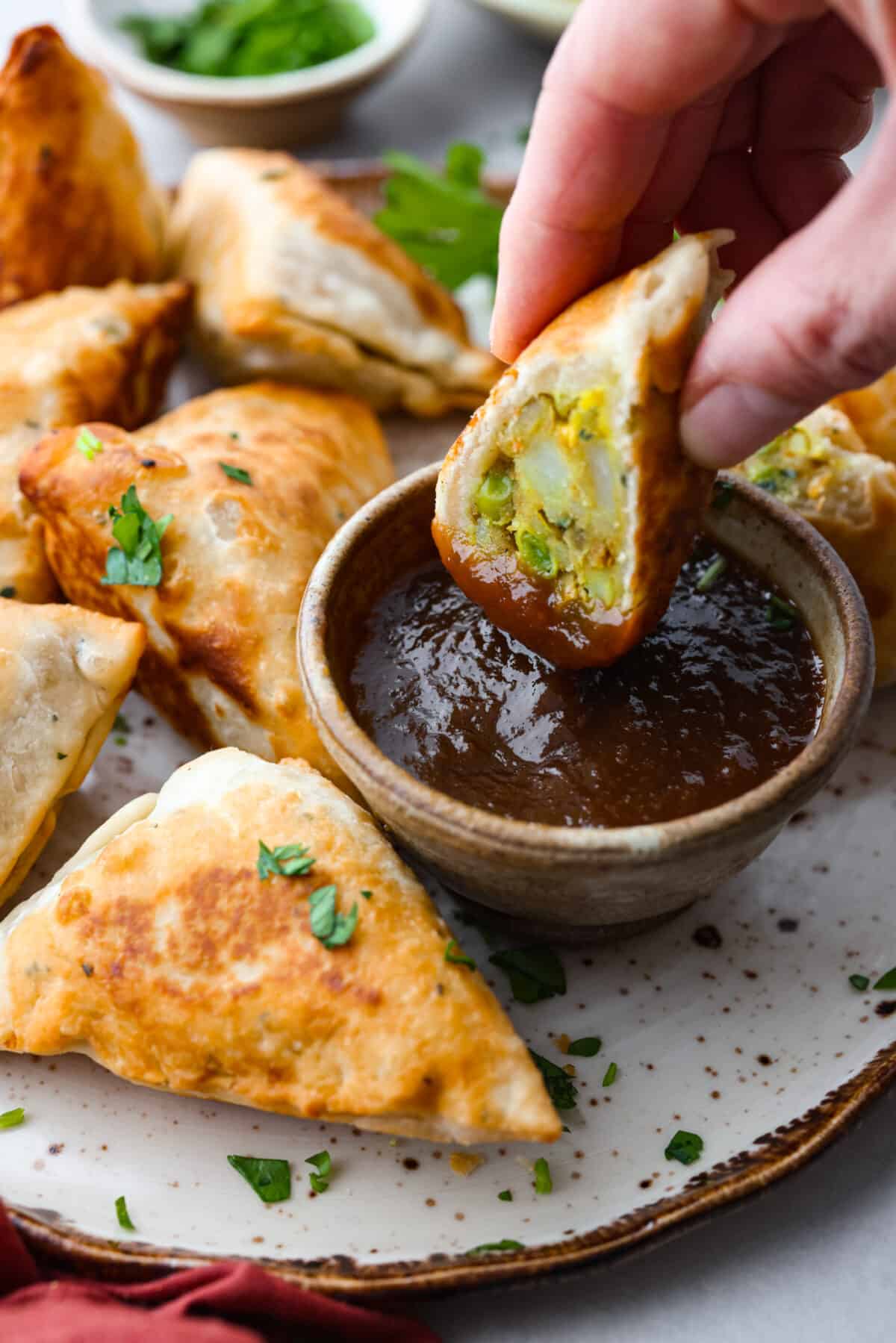 Craving some crispy, golden goodness? Look no further than these fried potato and pea samosas, seasoned to perfection with a savory curry blend. Dipping half of a samosa into a thick, red tamarind sauce.