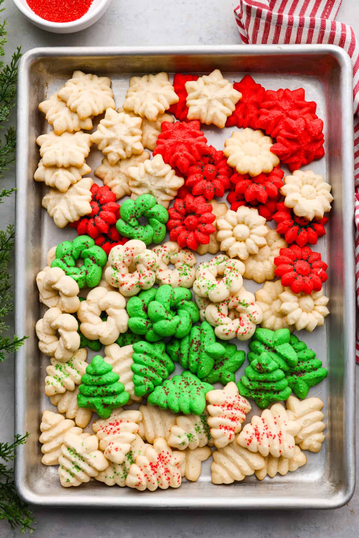 Spritz cookies are buttery and delicious, made with a cookie press to create festive shapes! Perfect for adding to any holiday dessert spread. Spritz cookies in a baking sheet. Some are dyed green or red. They are in the shape of Christmas trees and snowflakes.