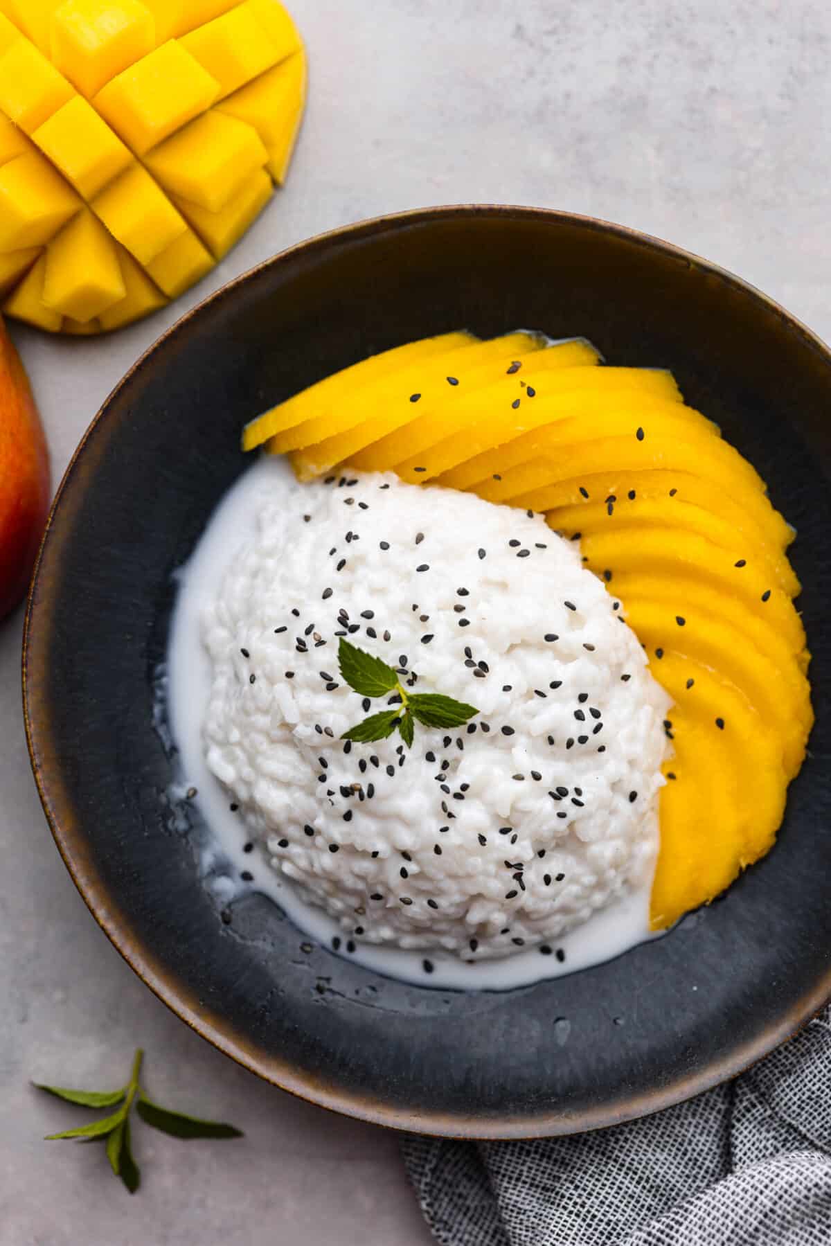Dive into this irresistible mango sticky rice! Warm coconut sauce, fluffy rice, and juicy mangoes - an iconic Thai dessert made with love in your own home. Top-down view of sticky rice served with coconut sauce and sliced mangoes.