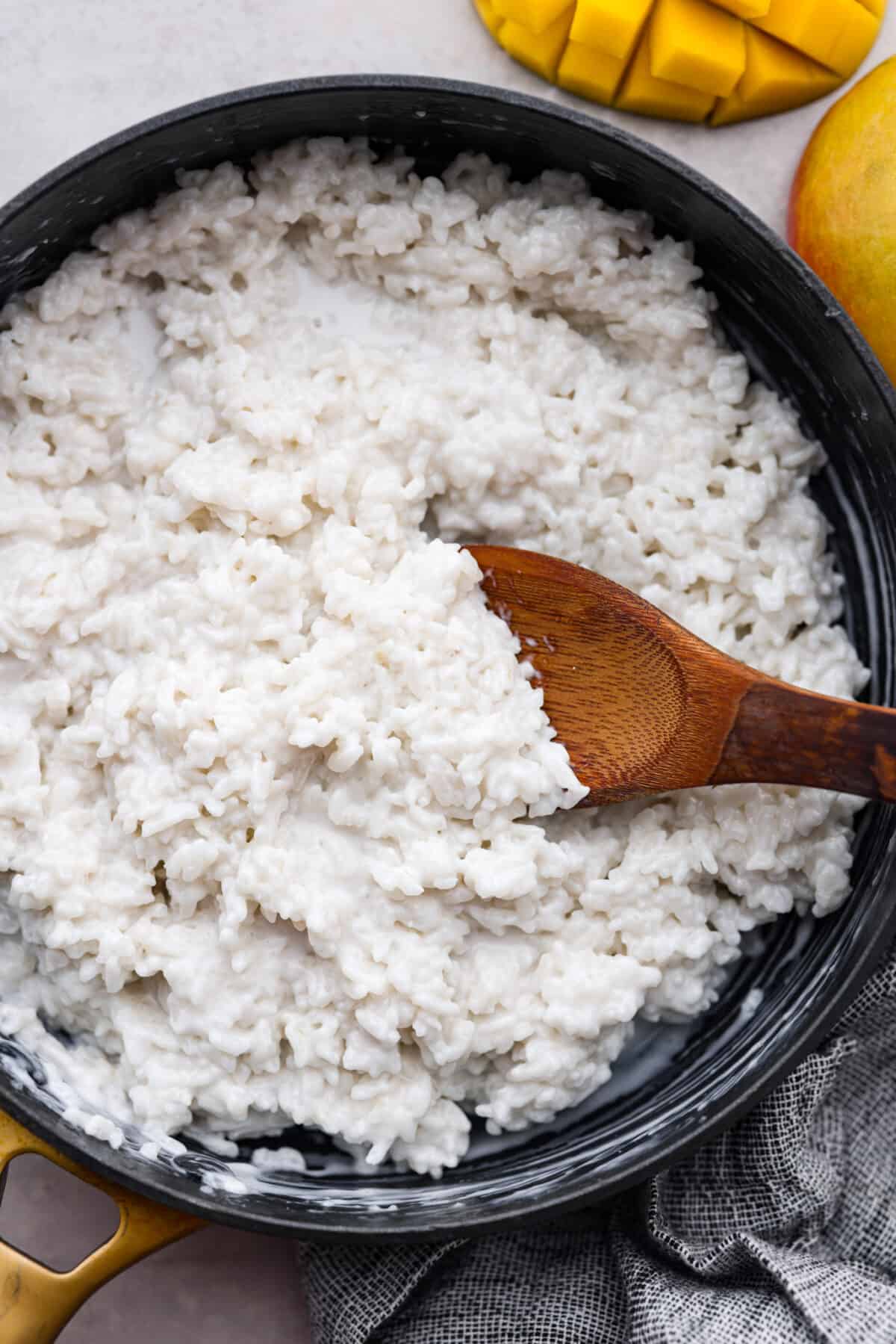 Dive into this irresistible mango sticky rice! Warm coconut sauce, fluffy rice, and juicy mangoes - an iconic Thai dessert made with love in your own home. Closeup of the sticky rice in a black pan.