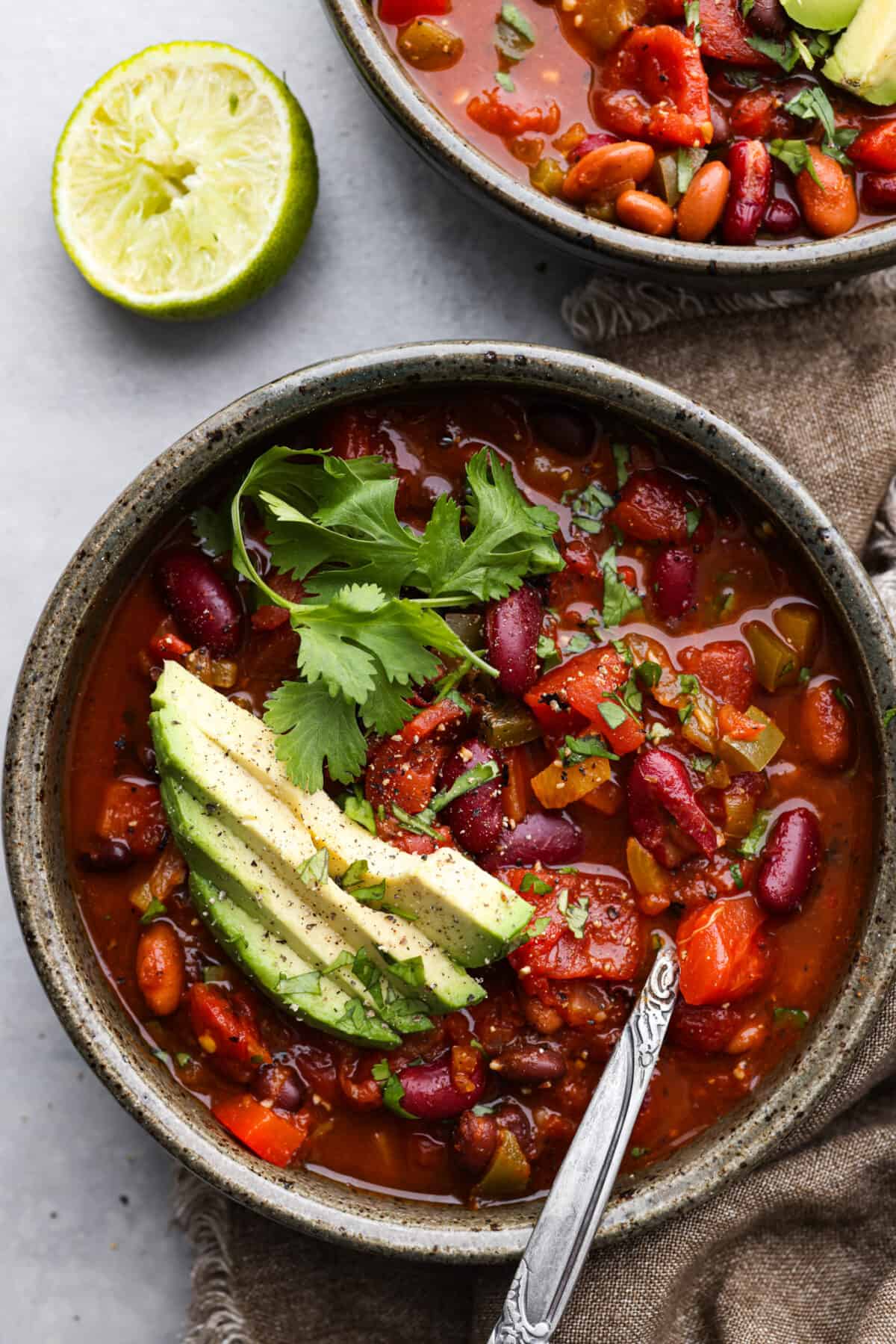 Introducing the ULTIMATE vegetarian chili recipe! It's full of tender vegetables, a combination of beans, and loaded with seasonings. It's an easy one-pot chili bursting with so many flavors and textures that you won't even miss the meat! Overhead view of chili in a bowl with a spoon. Avocado and cilantro are garnished on top.