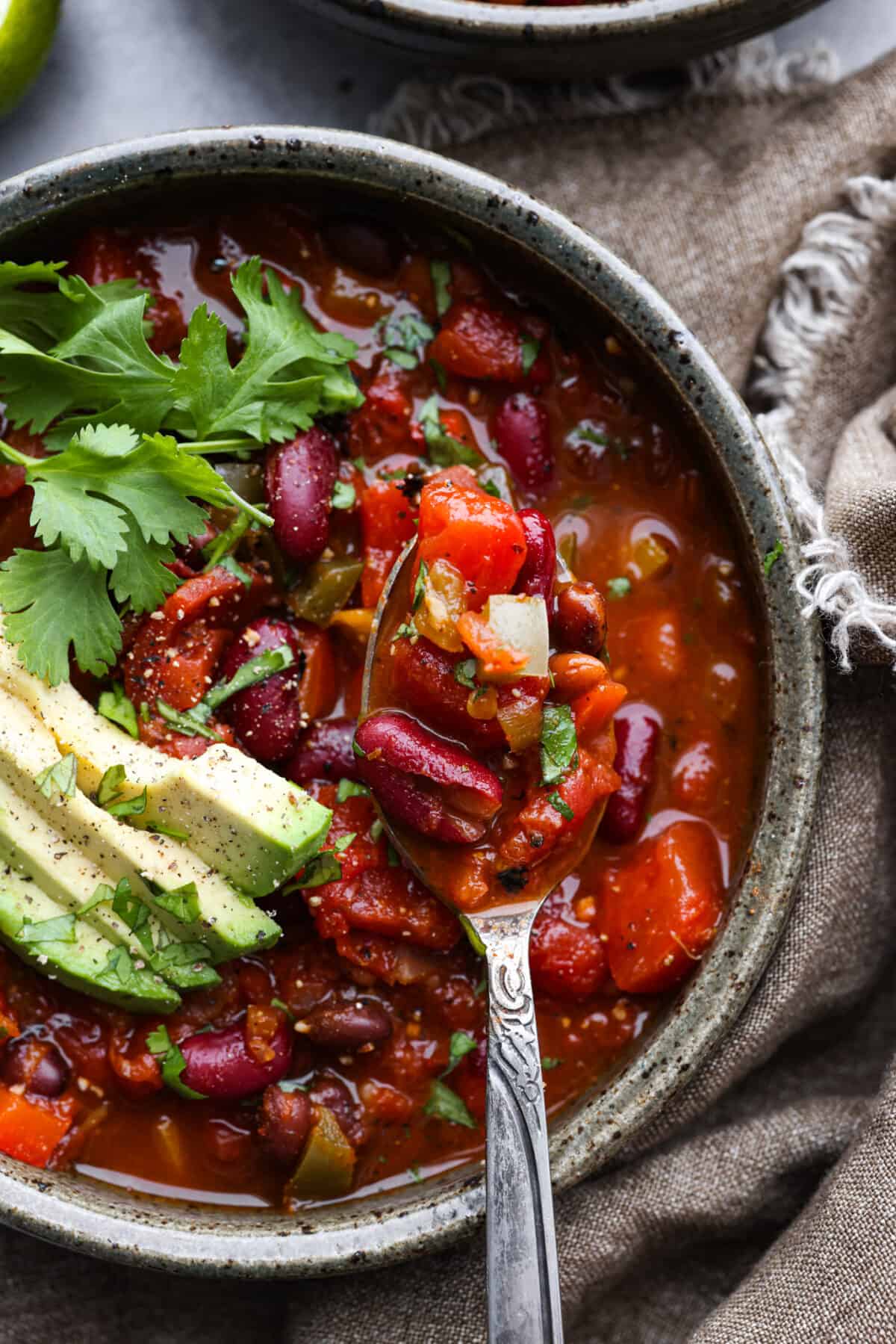 Introducing the ULTIMATE vegetarian chili recipe! It's full of tender vegetables, a combination of beans, and loaded with seasonings. It's an easy one-pot chili bursting with so many flavors and textures that you won't even miss the meat! Close up of chili in a gray bowl topped with cilantro and avocado.