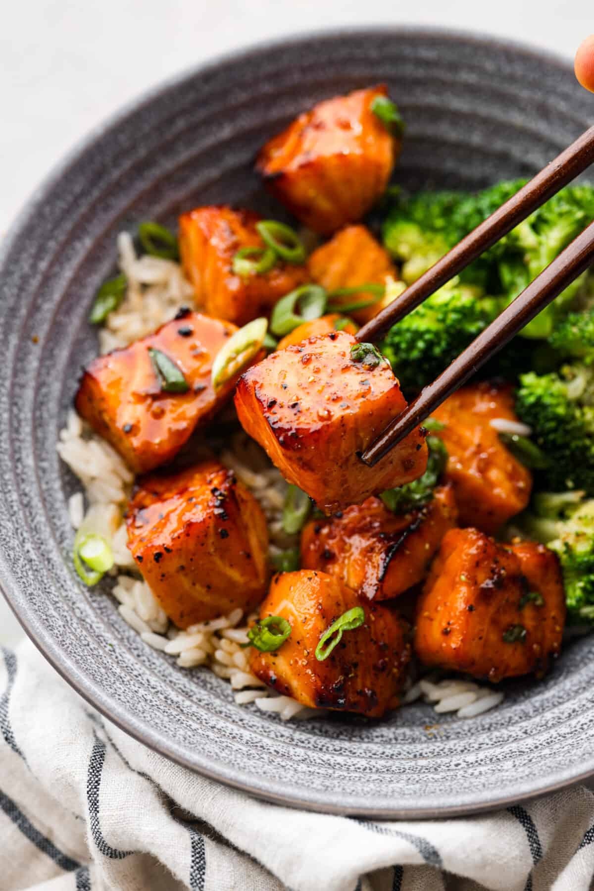 Cooked in less than 10 minutes, these perfectly glazed honey garlic air fryer salmon bites are so delicious. This is such a quick dinner, you will never want to cook salmon any other way! Close view of chopsticks lifting up a salmon bite in a bowl with rice and broccoli.