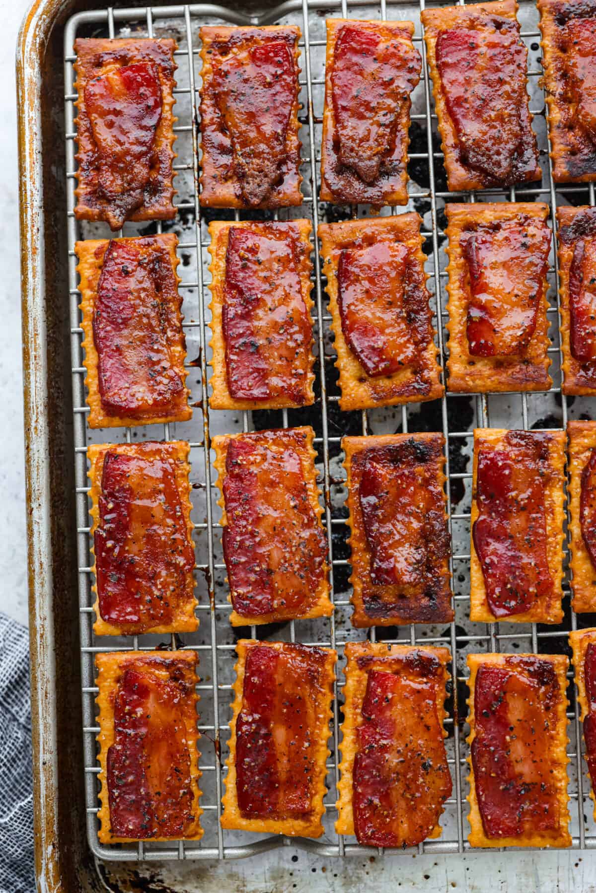 Introducing the easiest and tastiest appetizer that every party needs! Bacon crackers start with a buttery cracker layered with salty bacon and sprinkled with brown sugar and black pepper. It only takes 4 ingredients to make this sweet and salty snack. Close view of baked bacon crackers on a baking sheet pan lined with a wire rack.