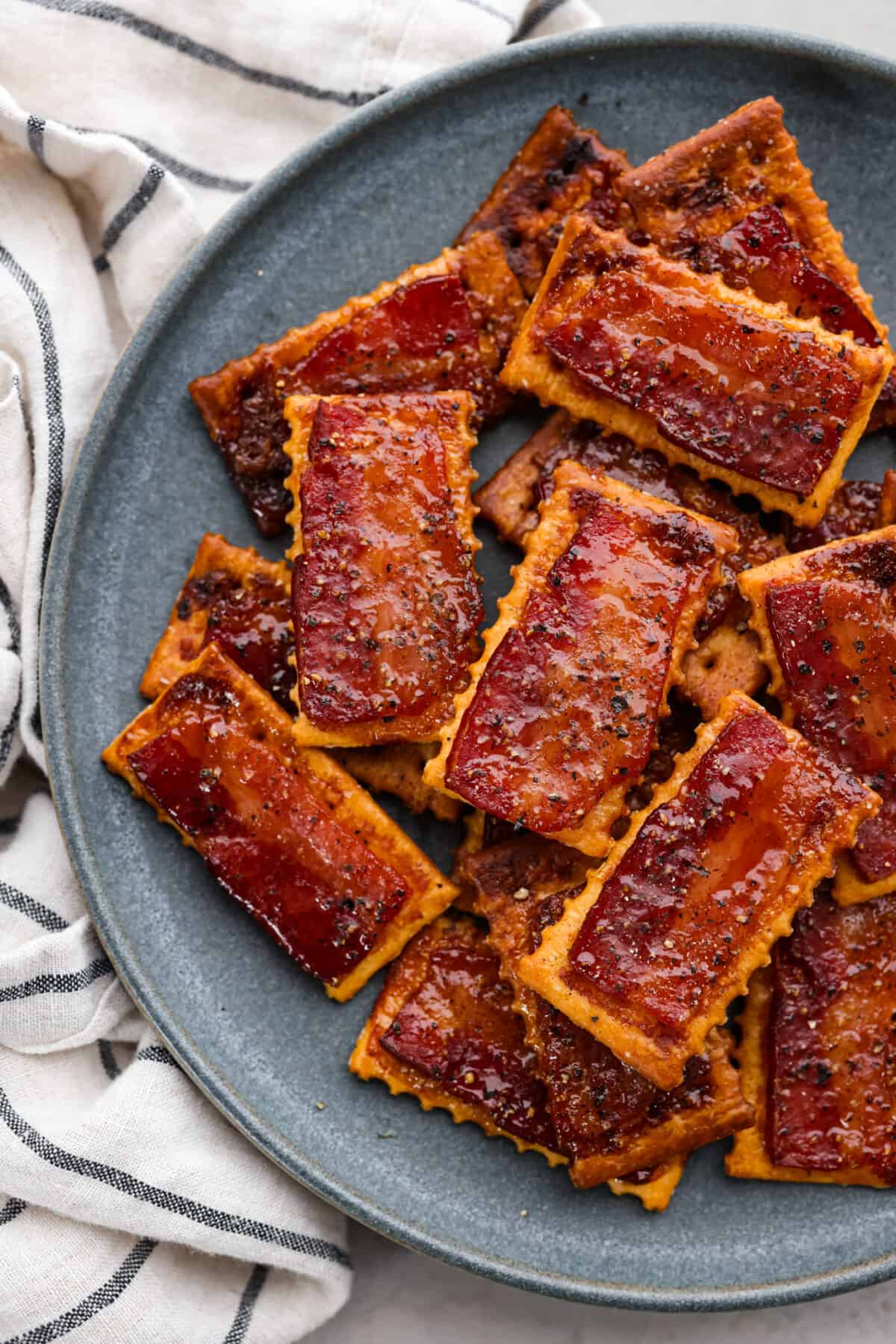 Introducing the easiest and tastiest appetizer that every party needs! Bacon crackers start with a buttery cracker layered with salty bacon and sprinkled with brown sugar and black pepper. It only takes 4 ingredients to make this sweet and salty snack. Close top view of bacon crackers stacked on a blue plate.