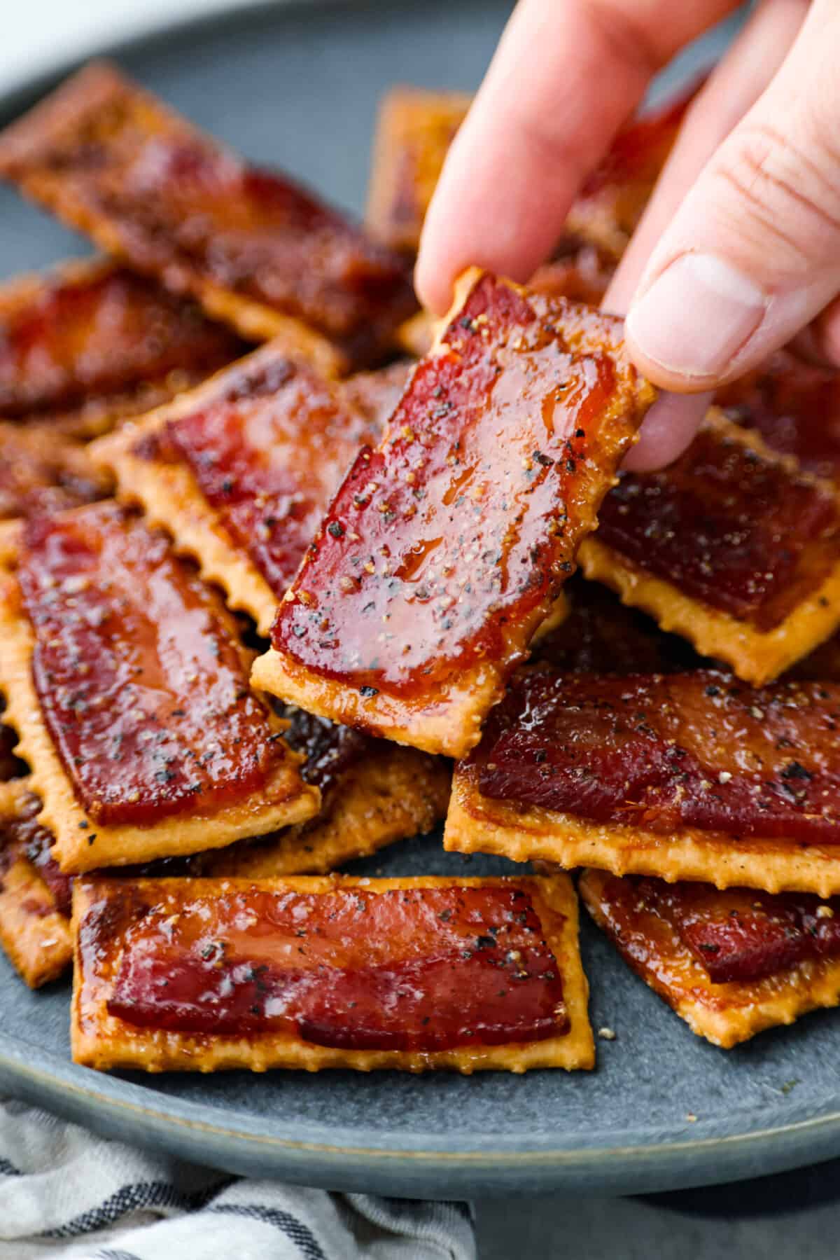 Introducing the easiest and tastiest appetizer that every party needs! Bacon crackers start with a buttery cracker layered with salty bacon and sprinkled with brown sugar and black pepper. It only takes 4 ingredients to make this sweet and salty snack. Close view of a hand holding up a bacon cracker from a plate of stacked crackers.