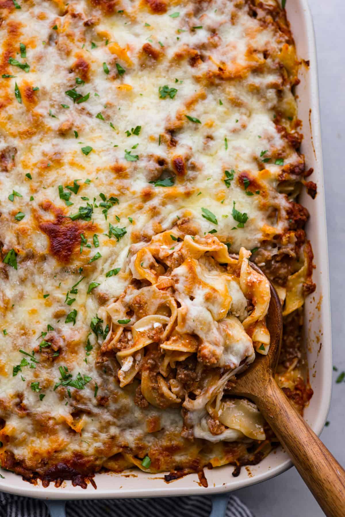 This flavorful Sour Cream Noodle Bake takes pasta to a whole new level! Using sour cream, cottage cheese, ground beef, tomato sauce, noodles and plenty of mozzarella, this recipe will have you coming back for seconds and even thirds! A scoop of sour cream noodle bake on a wooden spoon.
