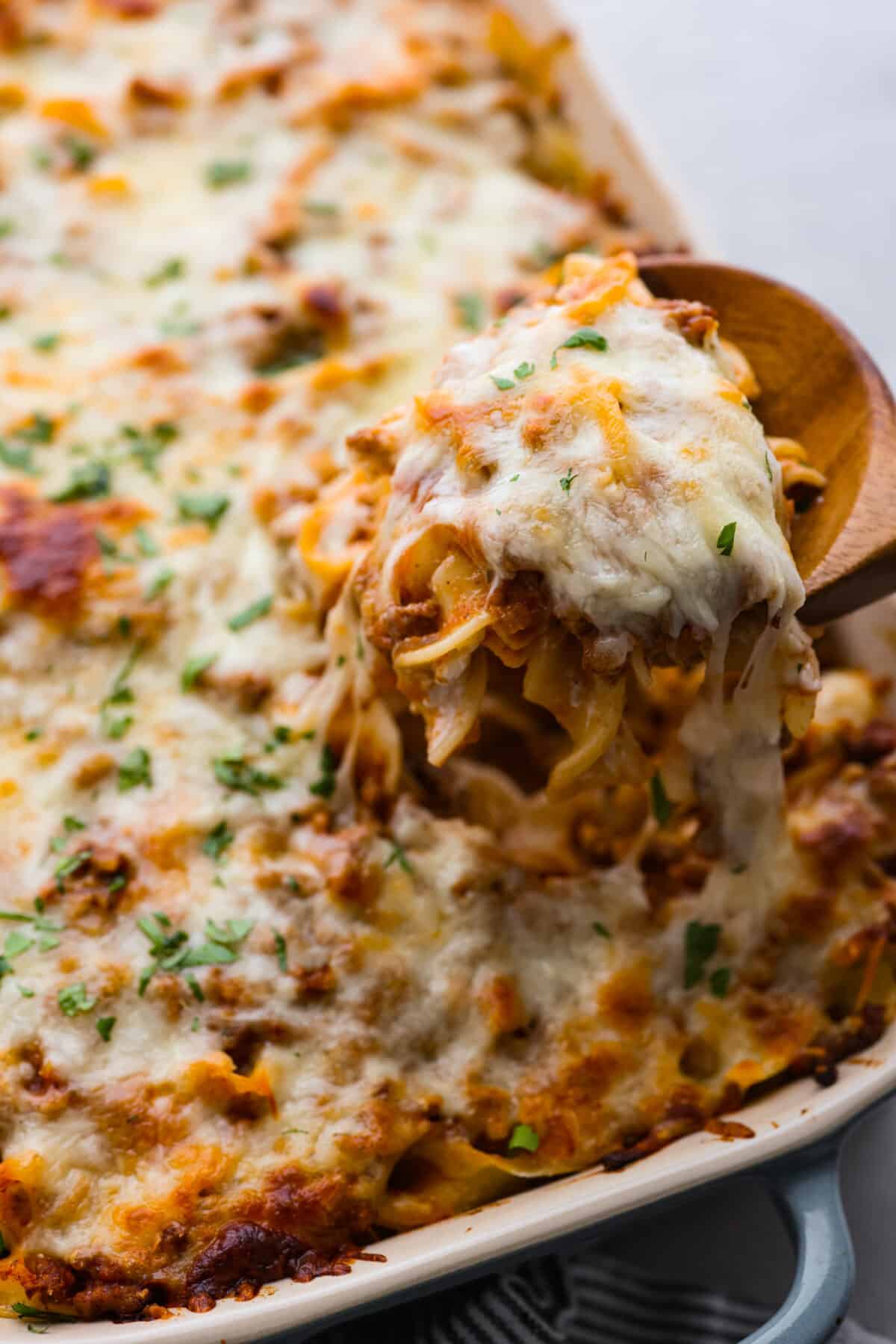 This flavorful Sour Cream Noodle Bake takes pasta to a whole new level! Using sour cream, cottage cheese, ground beef, tomato sauce, noodles and plenty of mozzarella, this recipe will have you coming back for seconds and even thirds! A cheesy scoop of sour cream noodle bake being pulled up with a wooden spoon.