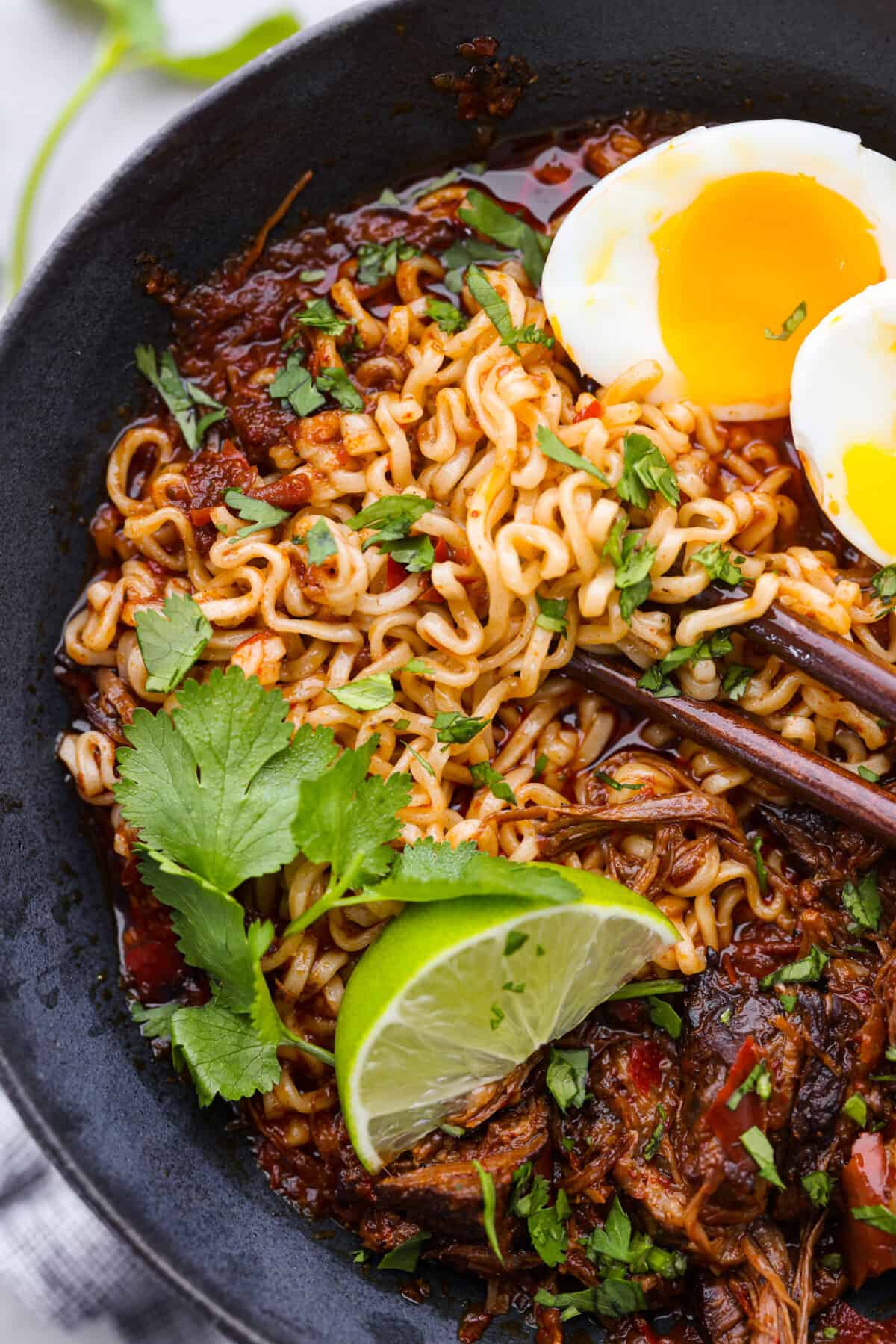 My Birria Ramen is next-level delicious! Tender shredded chuck roast, rich consommé, and slurp-worthy noodles all in one bowl. Closeup of noodles topped with cilantro and a lime wedge.