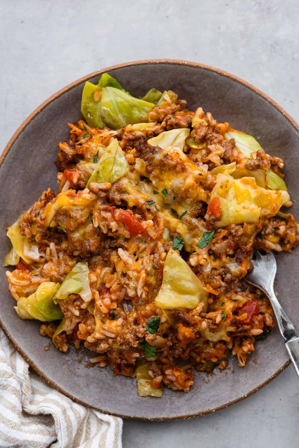 Cabbage roll casserole is a much EASIER version of cabbage rolls! Layers of ground beef, onion, garlic, rice, marinara, cheese, and tasty seasonings are assembled into a casserole. You'll love all the textures and flavors in this easy one-pan dinner! Top view of cabbage roll casserole on a brown plate with a fork.