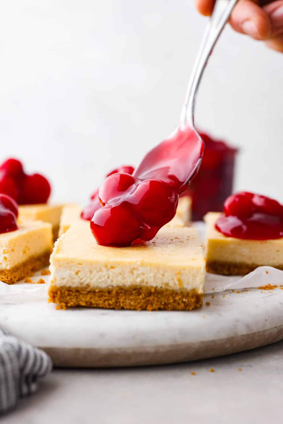 These Cherry Cheesecake Bars are the perfect make-ahead dessert for any occasion! Creamy cheesecake sits atop a homemade graham cracker crust, and is finished off with a generous helping of cherries. The cherry pie filling being added to a cheesecake bar.
