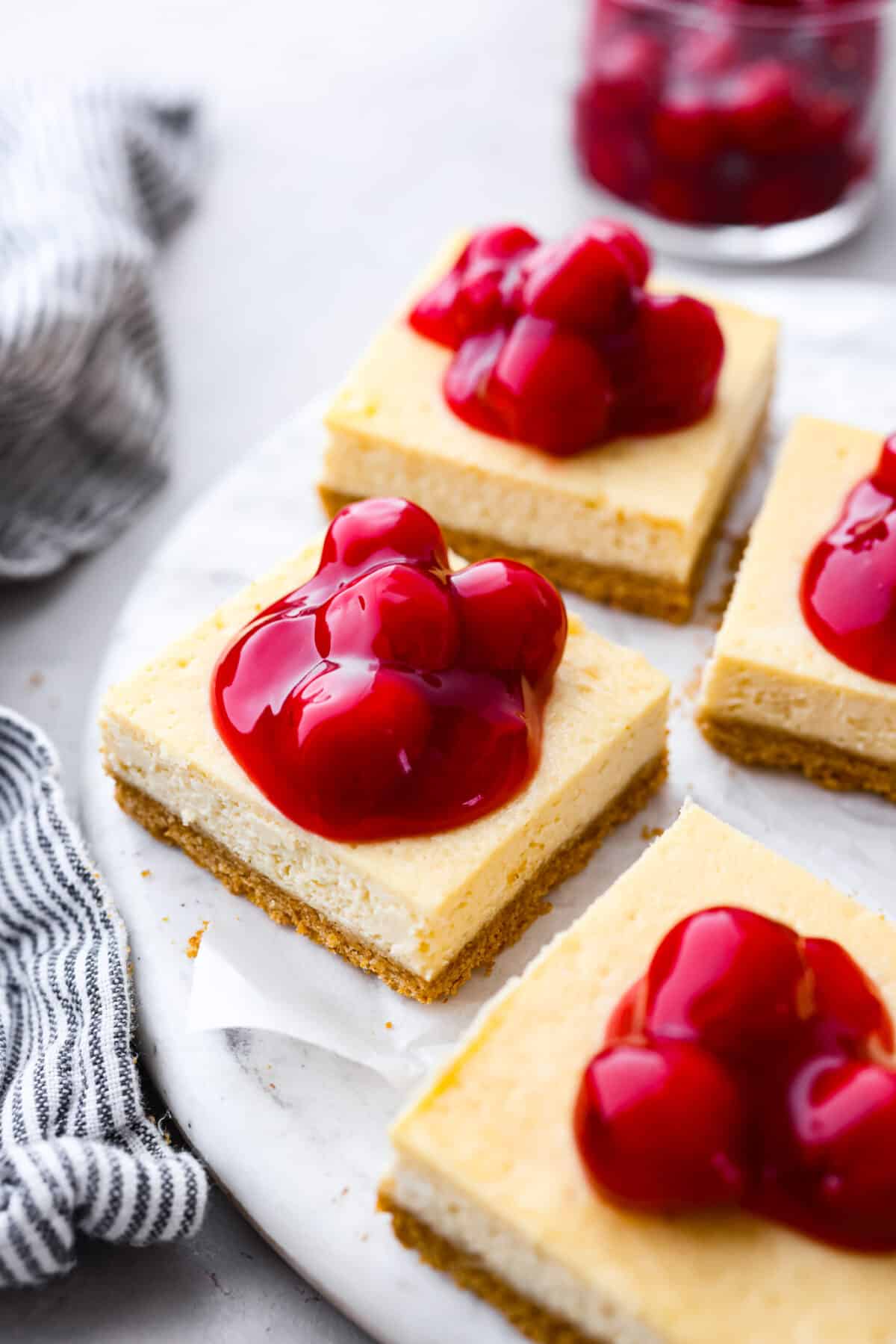 These Cherry Cheesecake Bars are the perfect make-ahead dessert for any occasion! Creamy cheesecake sits atop a homemade graham cracker crust, and is finished off with a generous helping of cherries. Sliced cheesecake bars topped with cherry pie filling on a white serving platter.