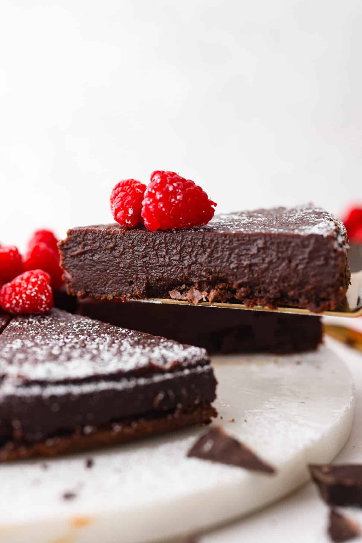 Flourless Chocolate Cake is an insanely rich and delicious chocolate dessert.ย Similar to the inside of a truffle, this gluten-free dessert will satisfy your sweet tooth with ease! A slice of chocolate cake being served with a cake spatula. There are raspberries on top of the cake.