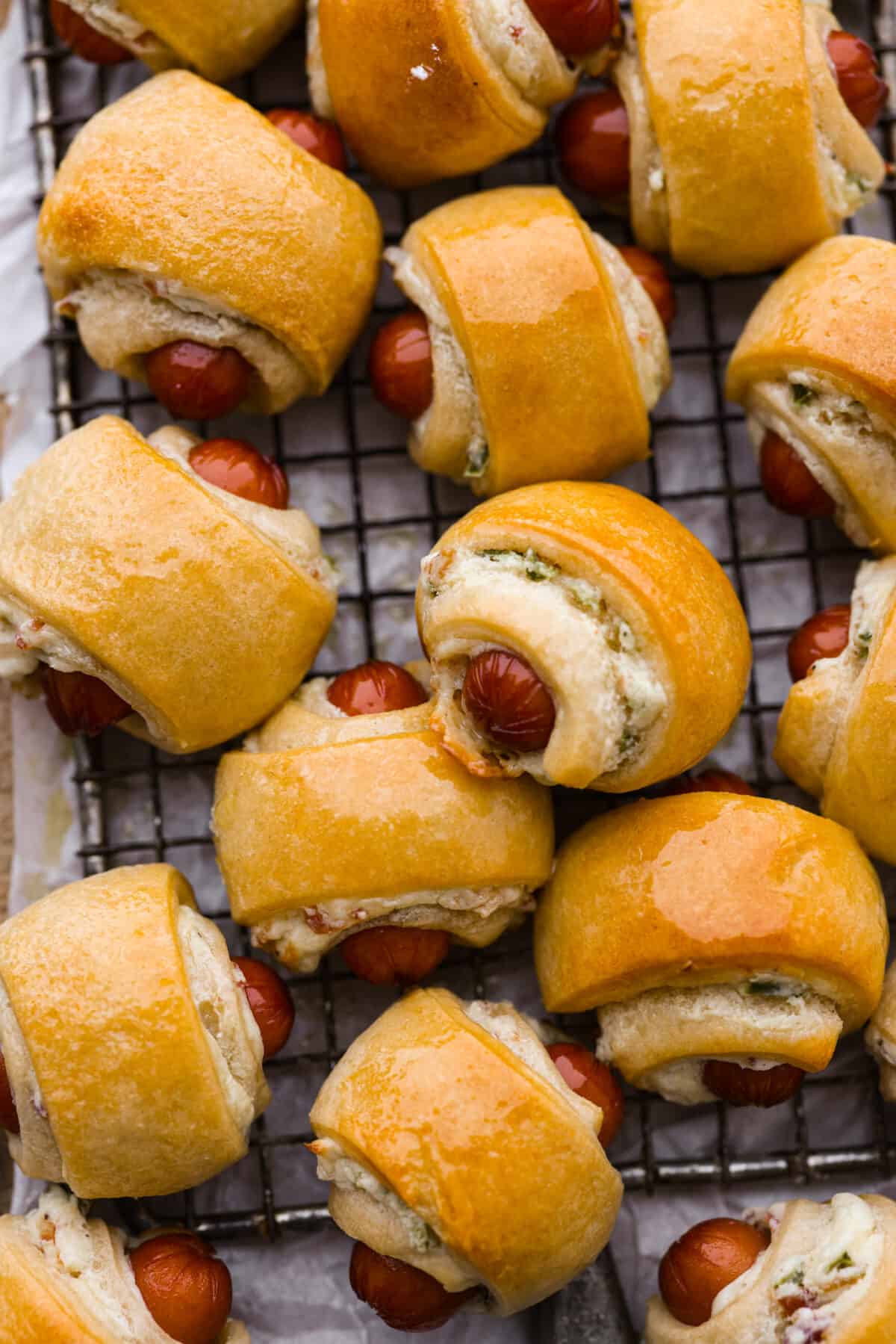 Upgrade the flavors of a classic appetizer with these jalapeño popper pigs in a blanket. Smokey cocktail sausages and jalapeño cream cheese wrapped in a blanket of buttery crescent dough. The jalapeño popper flavors add a creamy and spicy layer that tastes like it was meant to be there all along! Close view of pigs in a blanket close together on a cooling rack.