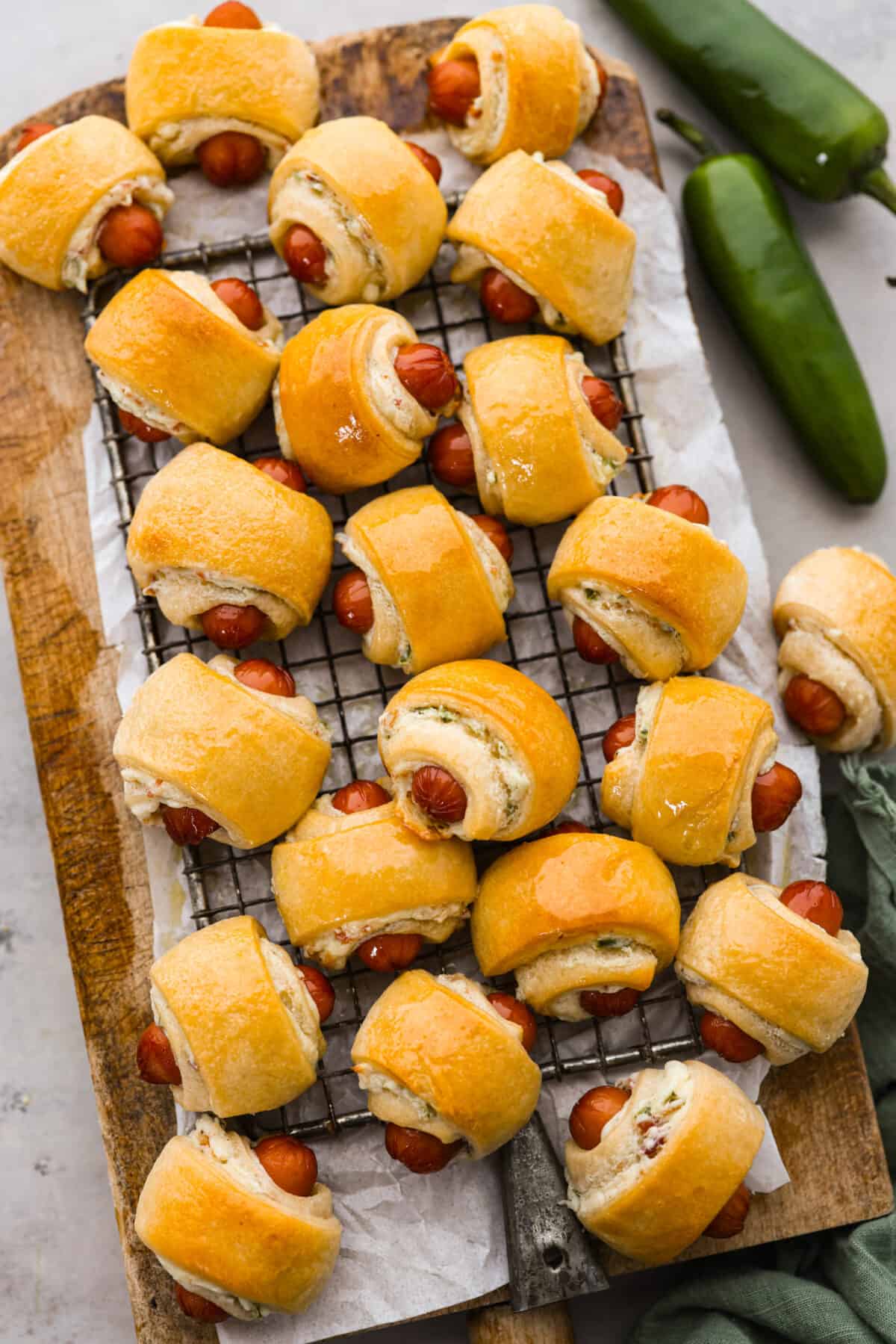 Upgrade the flavors of a classic appetizer with these jalapeño popper pigs in a blanket. Smokey cocktail sausages and jalapeño cream cheese wrapped in a blanket of buttery crescent dough. The jalapeño popper flavors add a creamy and spicy layer that tastes like it was meant to be there all along! Top view of jalapeno popper pigs in a blanket on a wood board lined with a cooling rack.