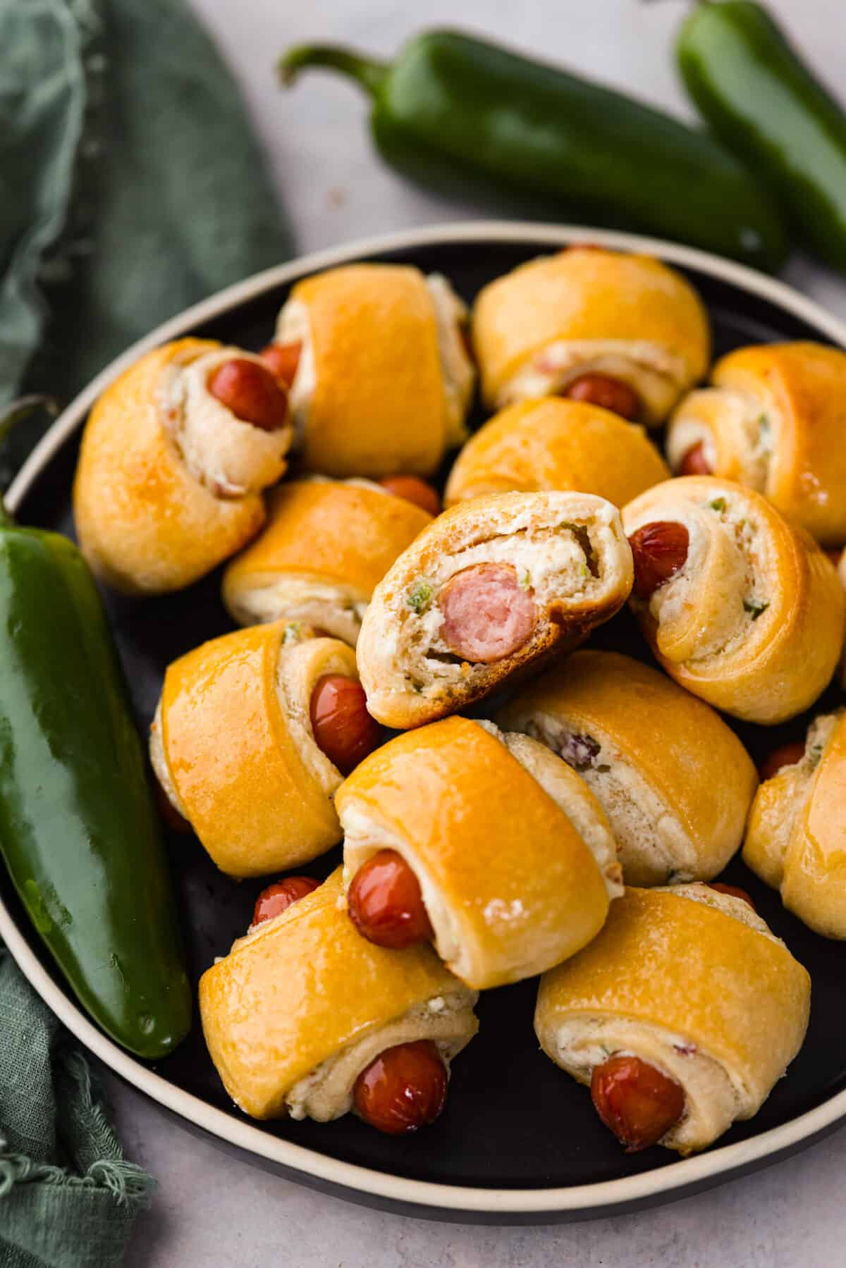 Upgrade the flavors of a classic appetizer with these jalapeño popper pigs in a blanket. Smokey cocktail sausages and jalapeño cream cheese wrapped in a blanket of buttery crescent dough. The jalapeño popper flavors add a creamy and spicy layer that tastes like it was meant to be there all along! Close view of jalapeno popper pigs in a blanket stacked on a black plate with a bite out of one.