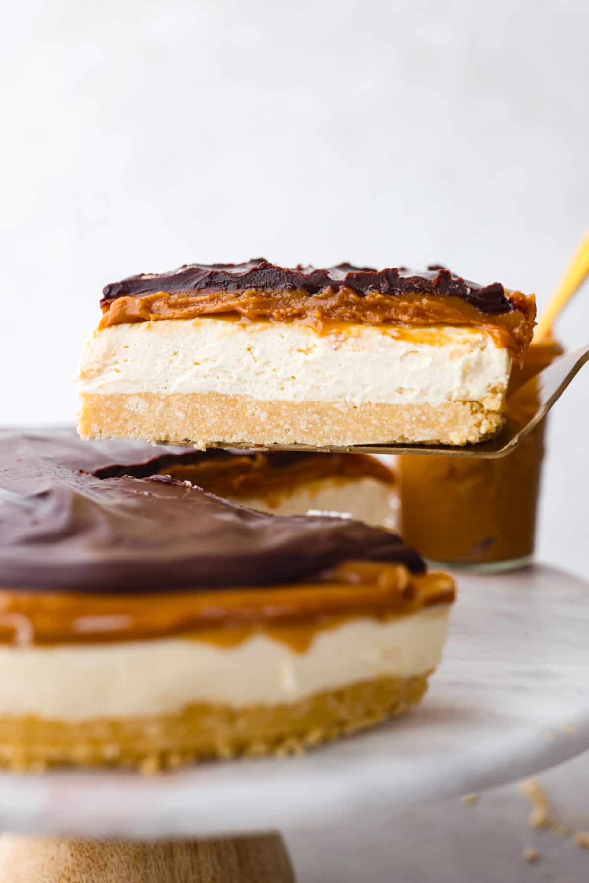 Delight in pure dessert heaven with this No-Bake Millionaire Cheesecake! It has four amazing layers that start with a shortbread crust, topped with a creamy cheesecake filling, then rich caramel and chocolate ganache to finish it off. And the best part? Itโs so easy to make with no baking required! Serving a slice of cheesecake with a cake spatula.